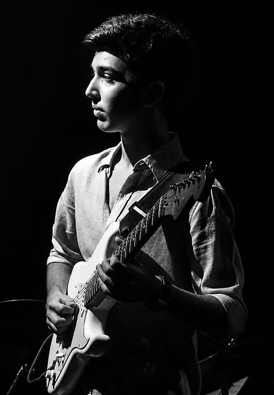 Person with short hair holding an electric guitar, wearing a shirt, in black and white, side profile.