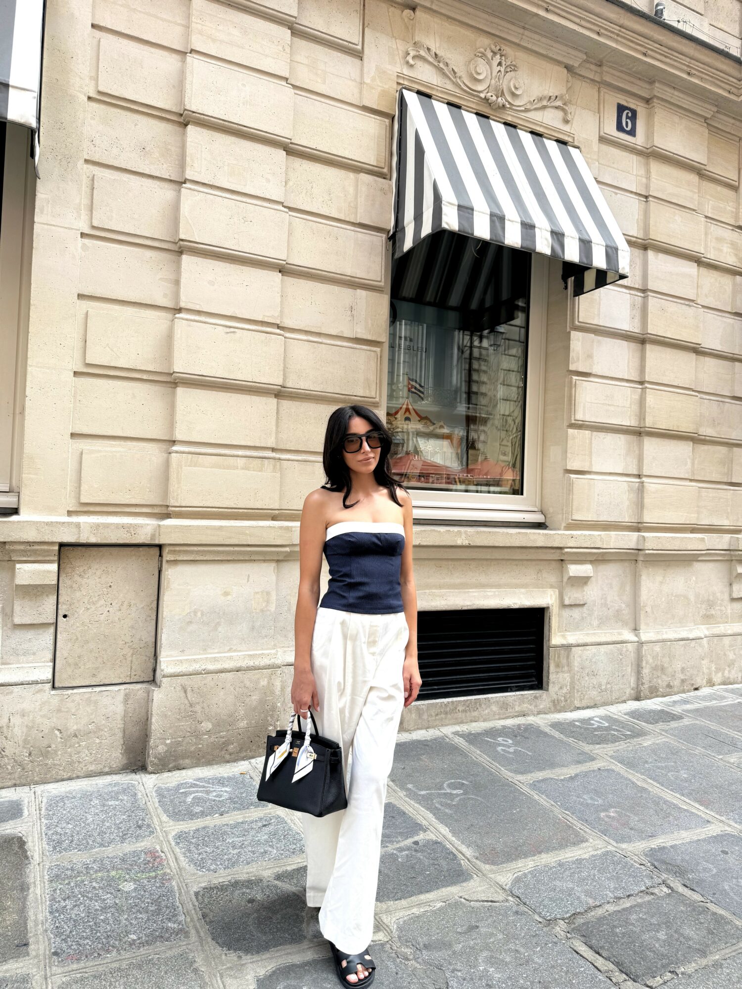 Woman in sunglasses standing outside a building with striped awning, holding a black and white bag, on a stone-paved street.