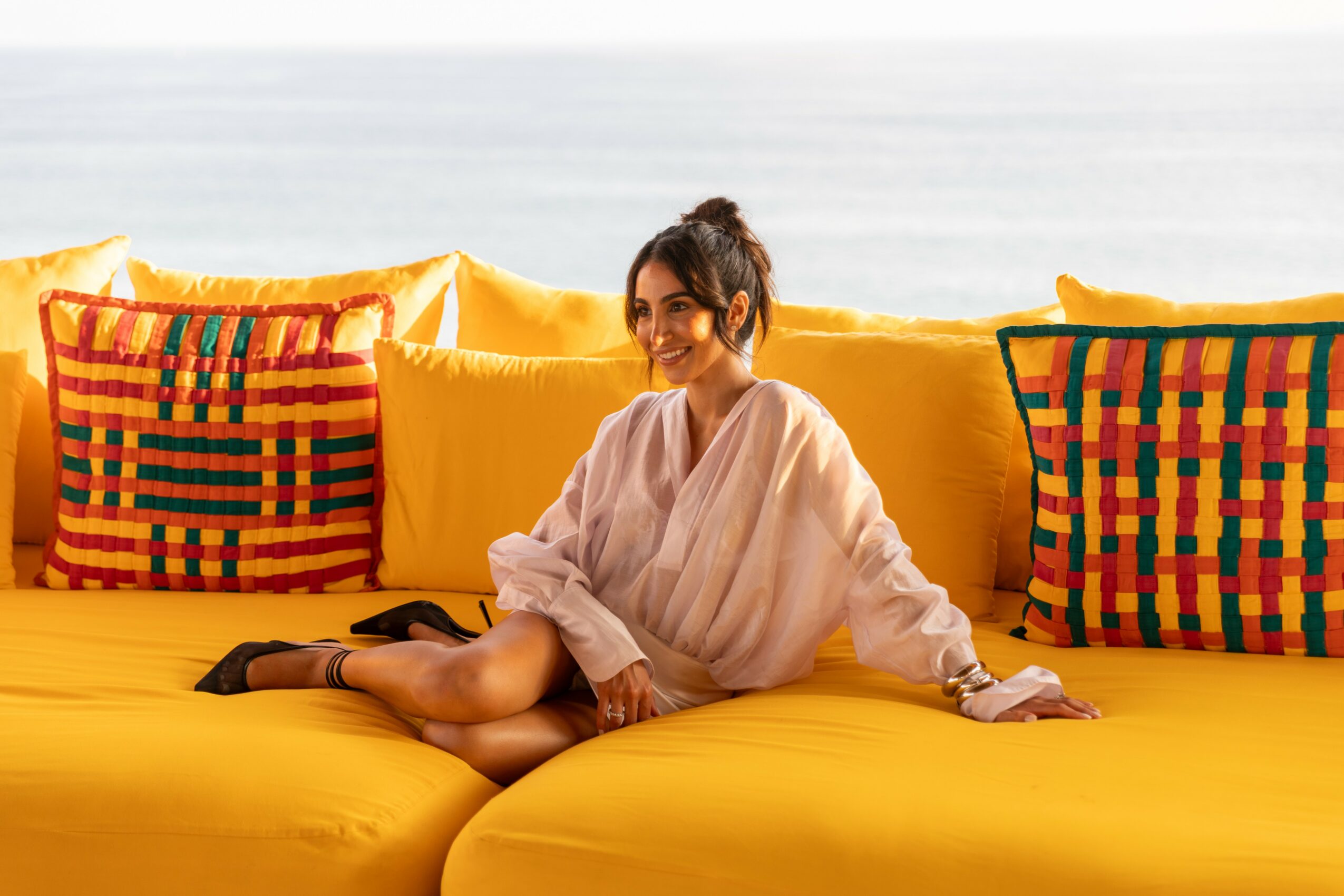 Woman sitting on yellow outdoor sofa with colorful cushions, smiling, with ocean in background.