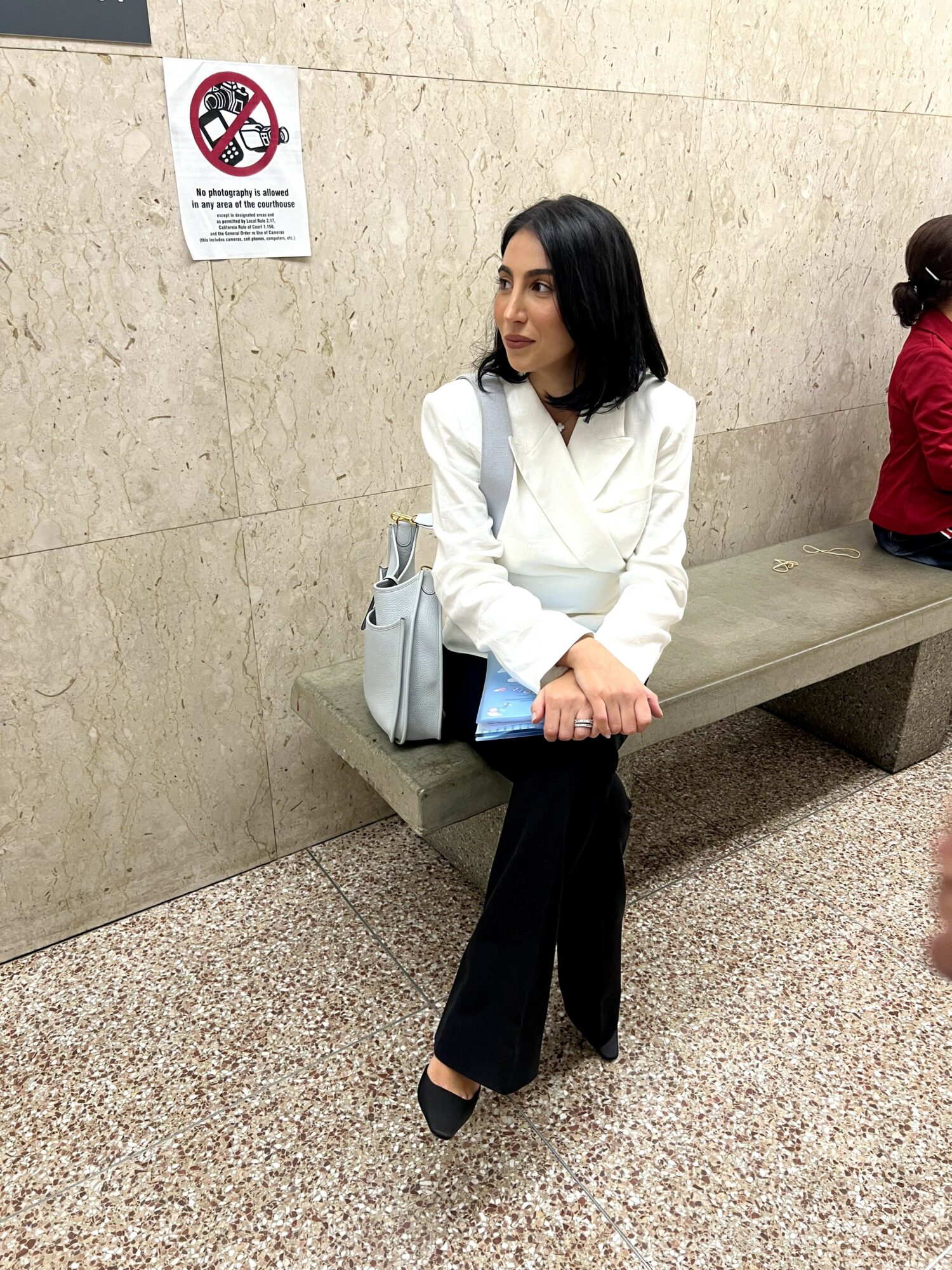 Woman sitting on a bench in a public space, wearing a white top and black pants, with a light-colored bag beside her.