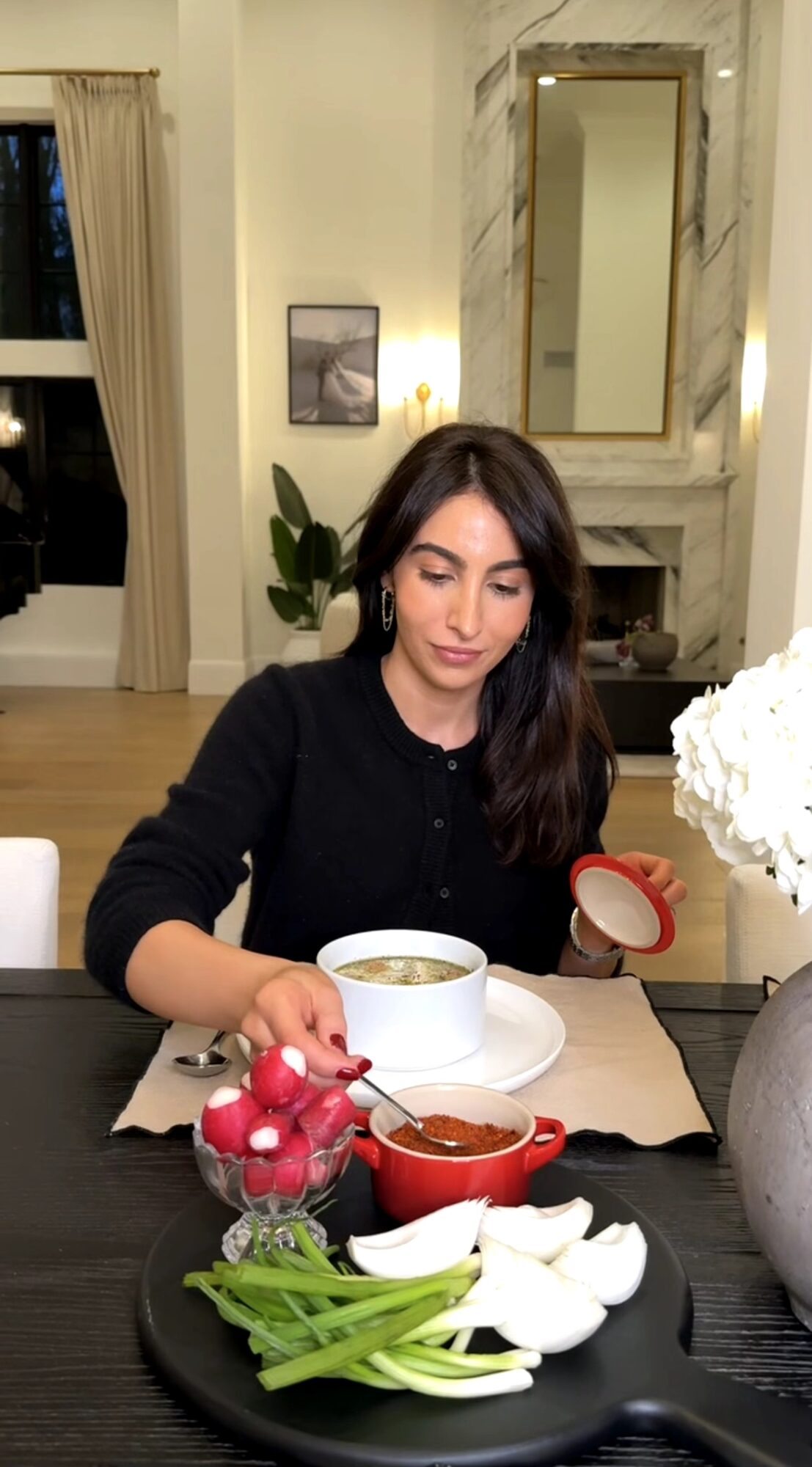 Young woman with dark hair in a black top sitting at a table with food and a cup, in a well-lit room with a mirror and plant.