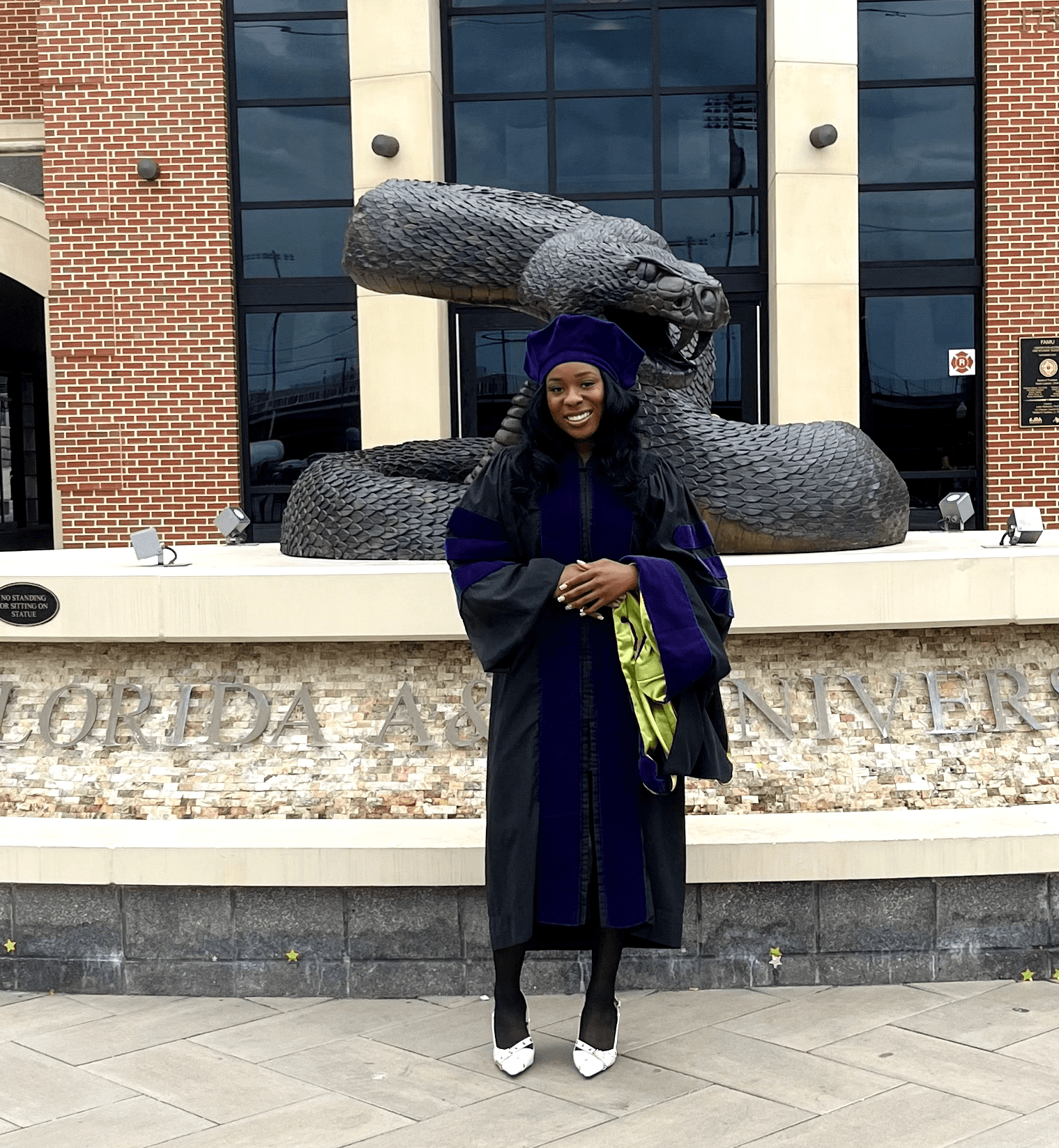 Person in graduation gown and cap standing in front of a snake sculpture outside a building.