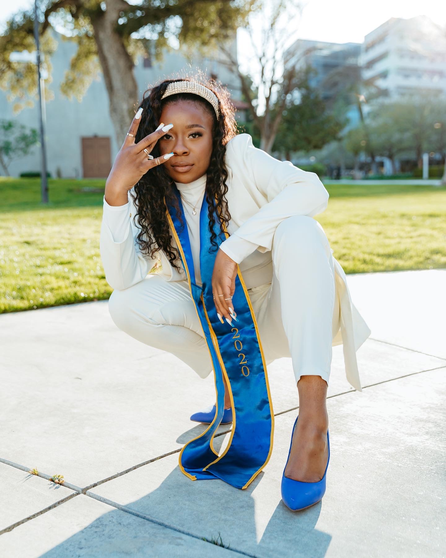 Young woman crouching outdoors, wearing white outfit, blue sash, and blue high heels, with headphones and making a peace sign gesture.