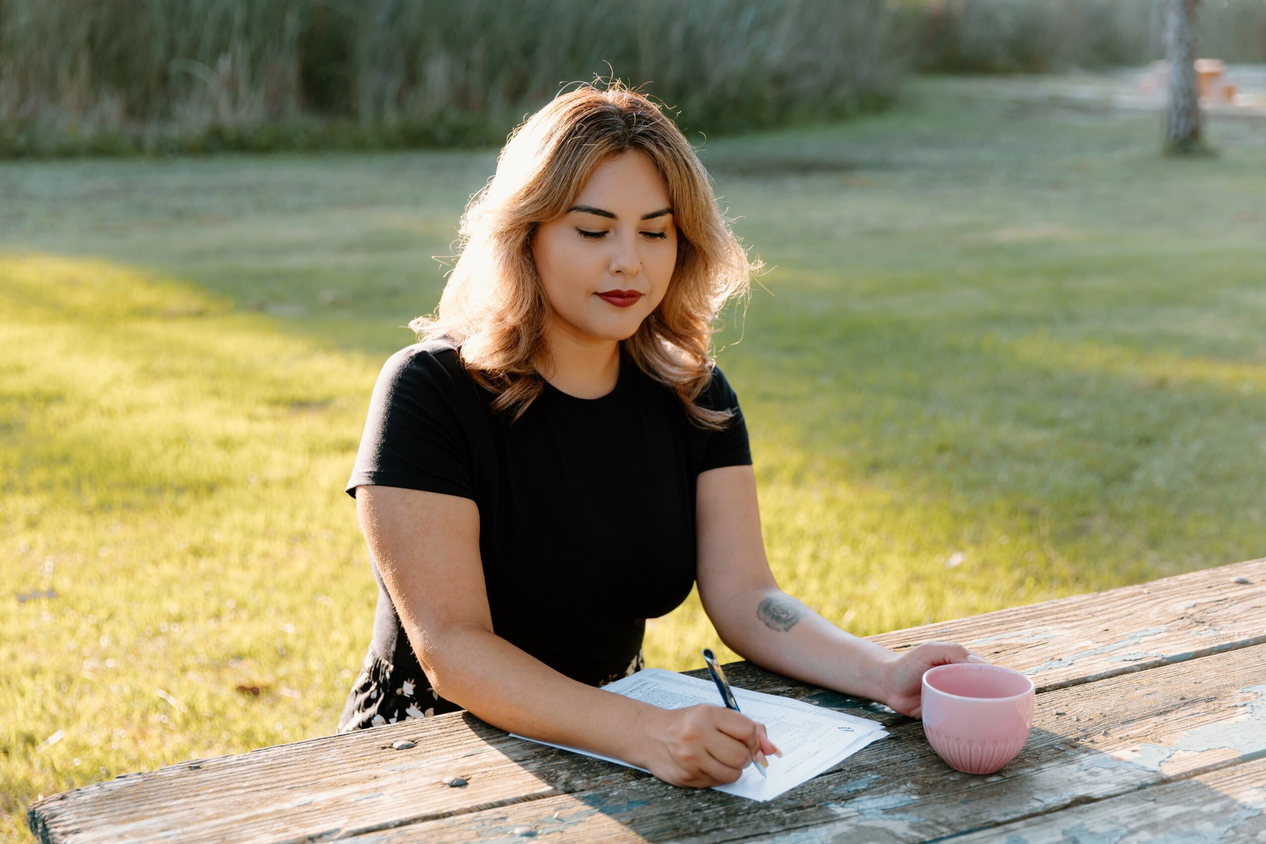Young woman with blonde hair sitting at a wooden table outdoors, writing on paper with a pink cup nearby.