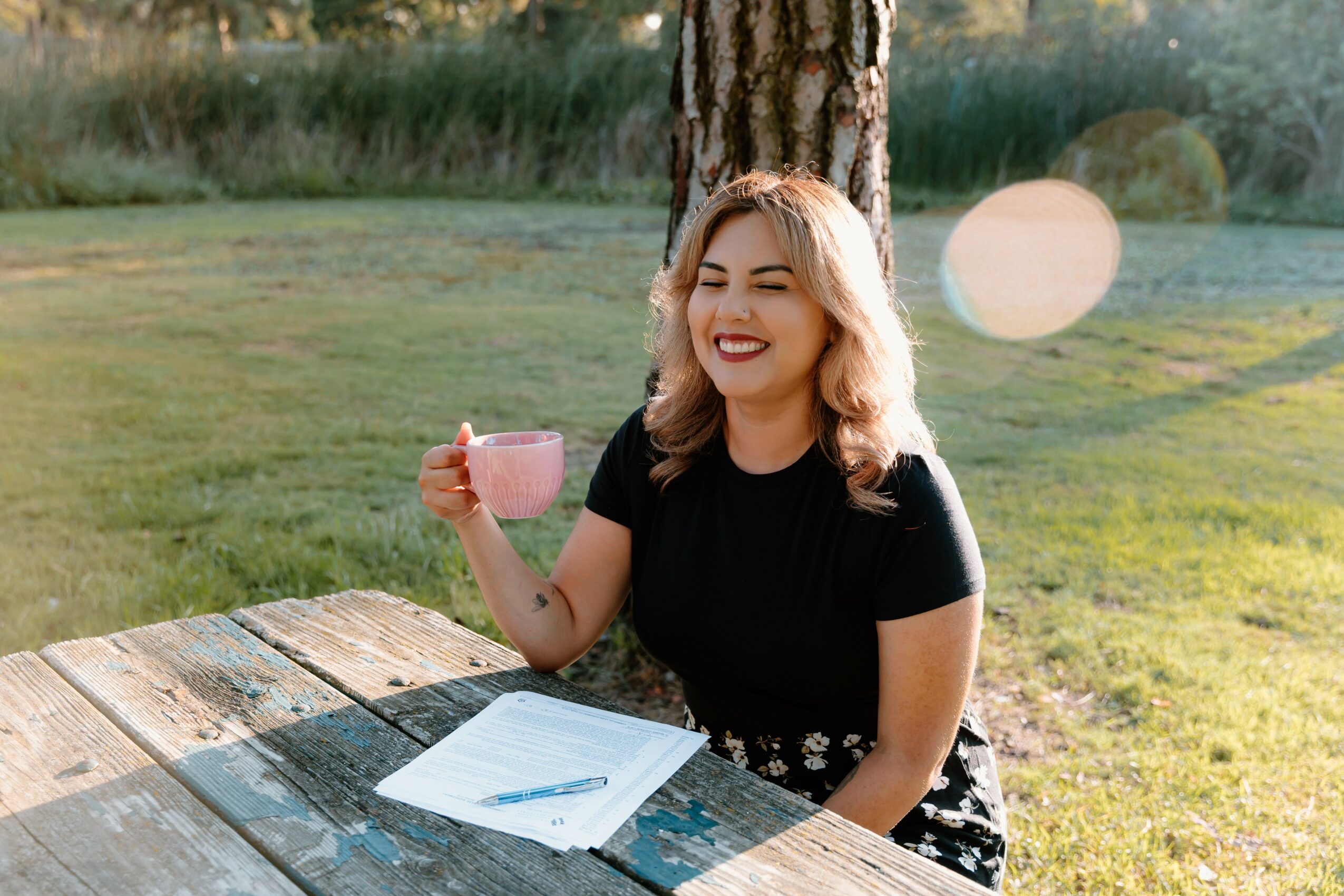 Young woman with shoulder-length hair smiling, holding a pink mug, sitting outdoors at a wooden table with papers, in sunlight.