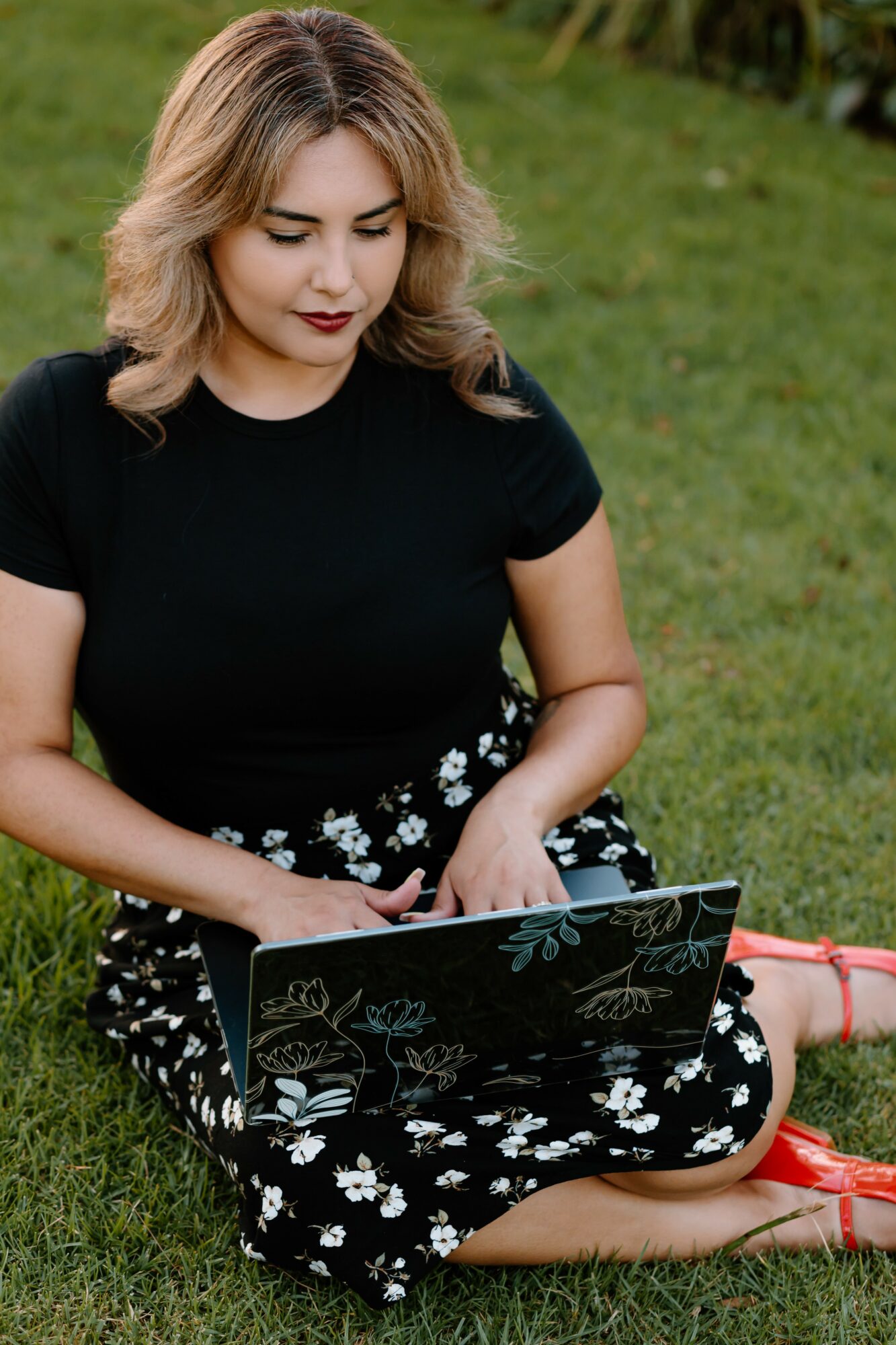 Woman sitting on grass with a laptop, wearing a black top and floral skirt, outdoors.
