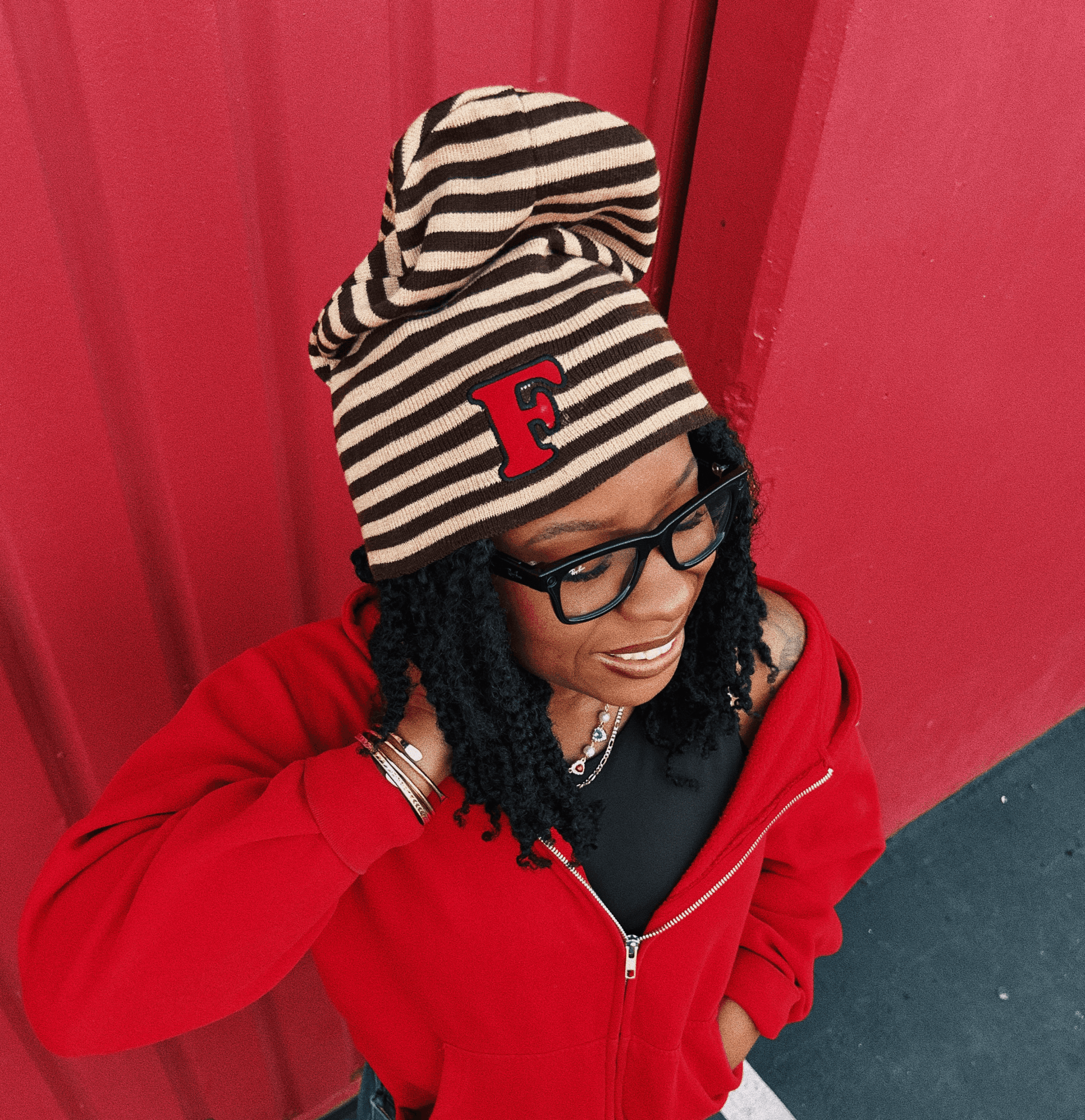 Person wearing a striped beanie with a red letter F, glasses, red jacket, and black hair, standing against a red wall.