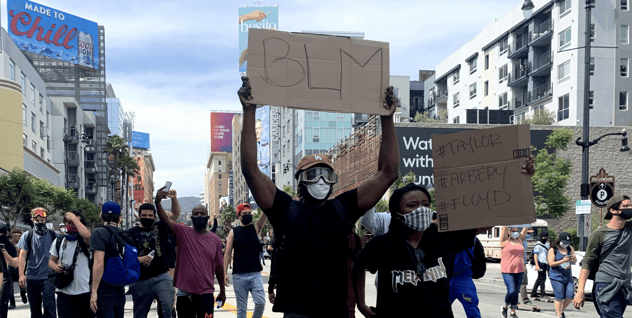 People protesting in a city street holding signs, with buildings and billboards in the background.