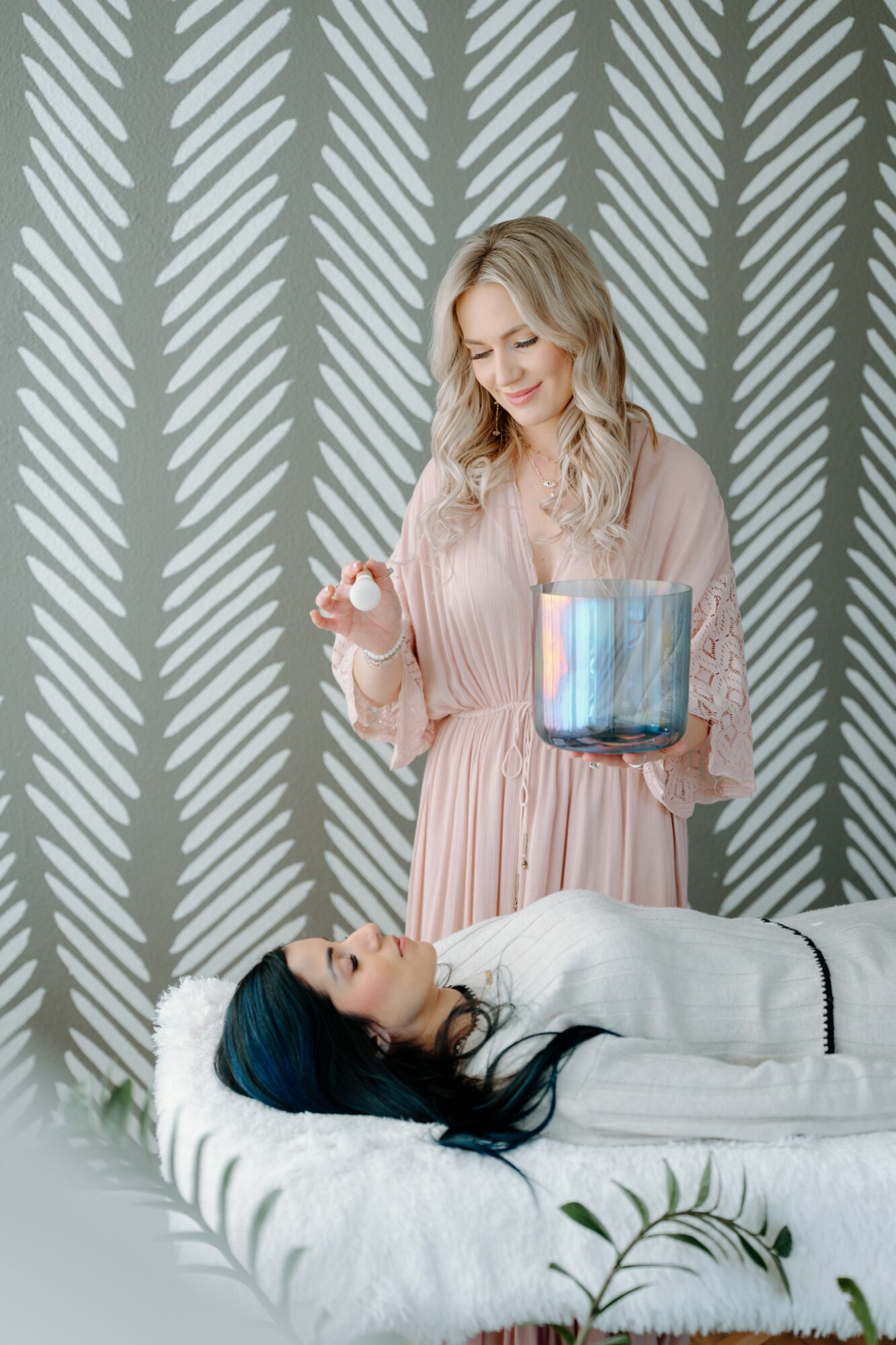 A woman in a pink dress holds a glass container with blue liquid, standing beside a person lying on a bed.