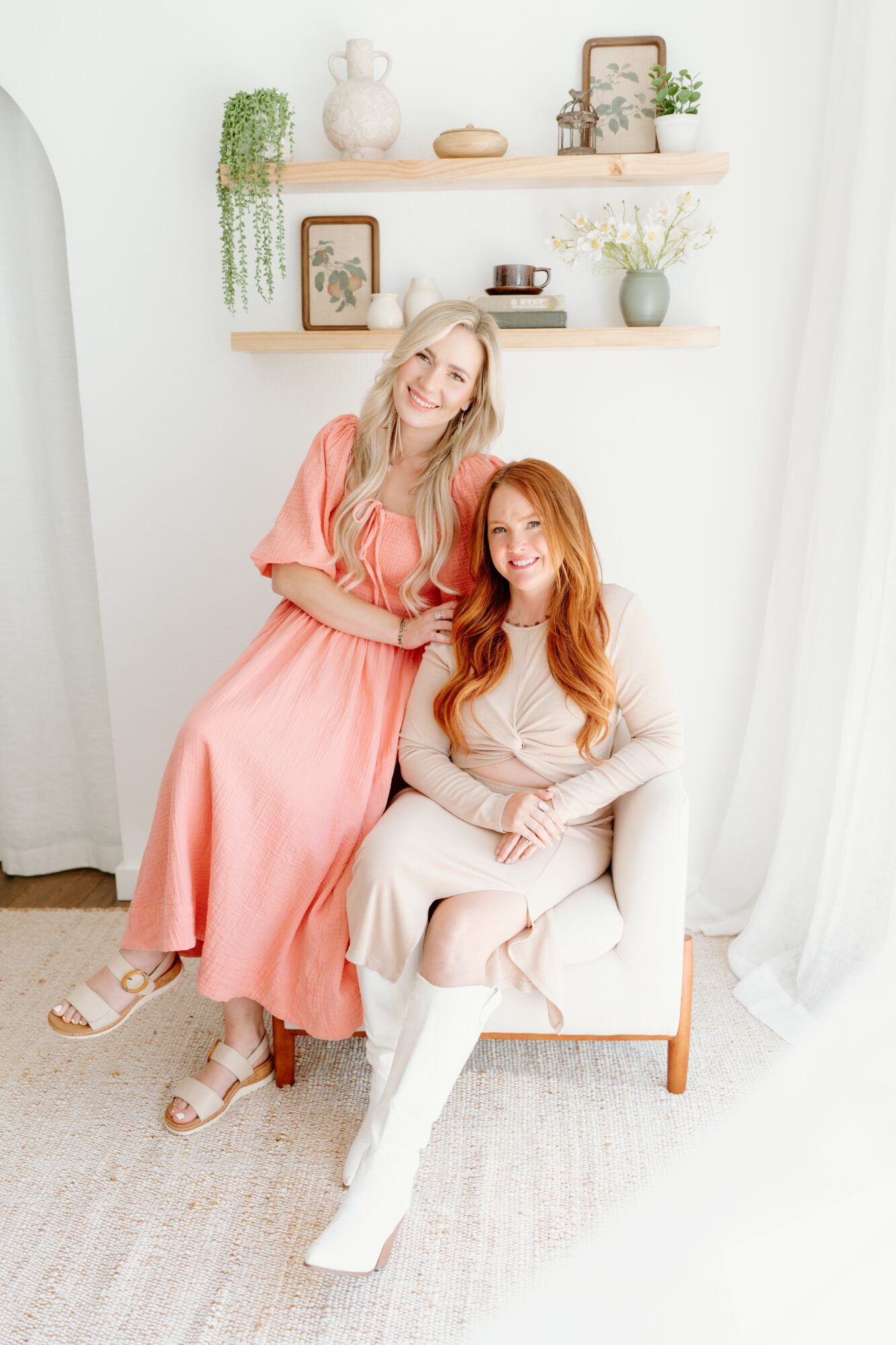 Two women with long hair sitting on a white chair in a bright room, smiling, with shelves and plants behind them.