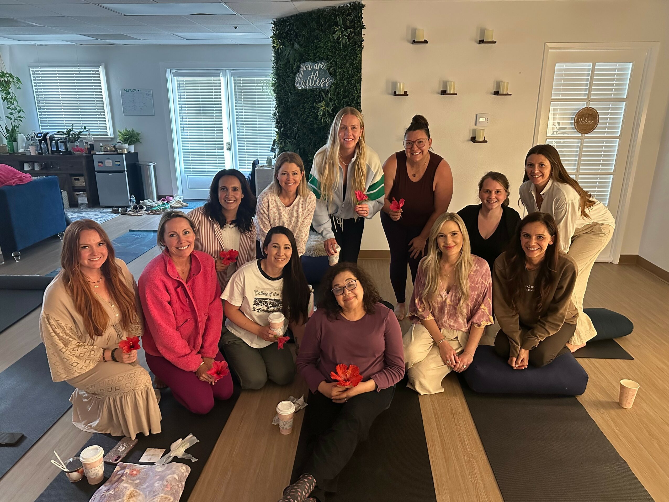 Group of twelve women sitting and standing in a living room, smiling, with a green wall and shelves behind them.