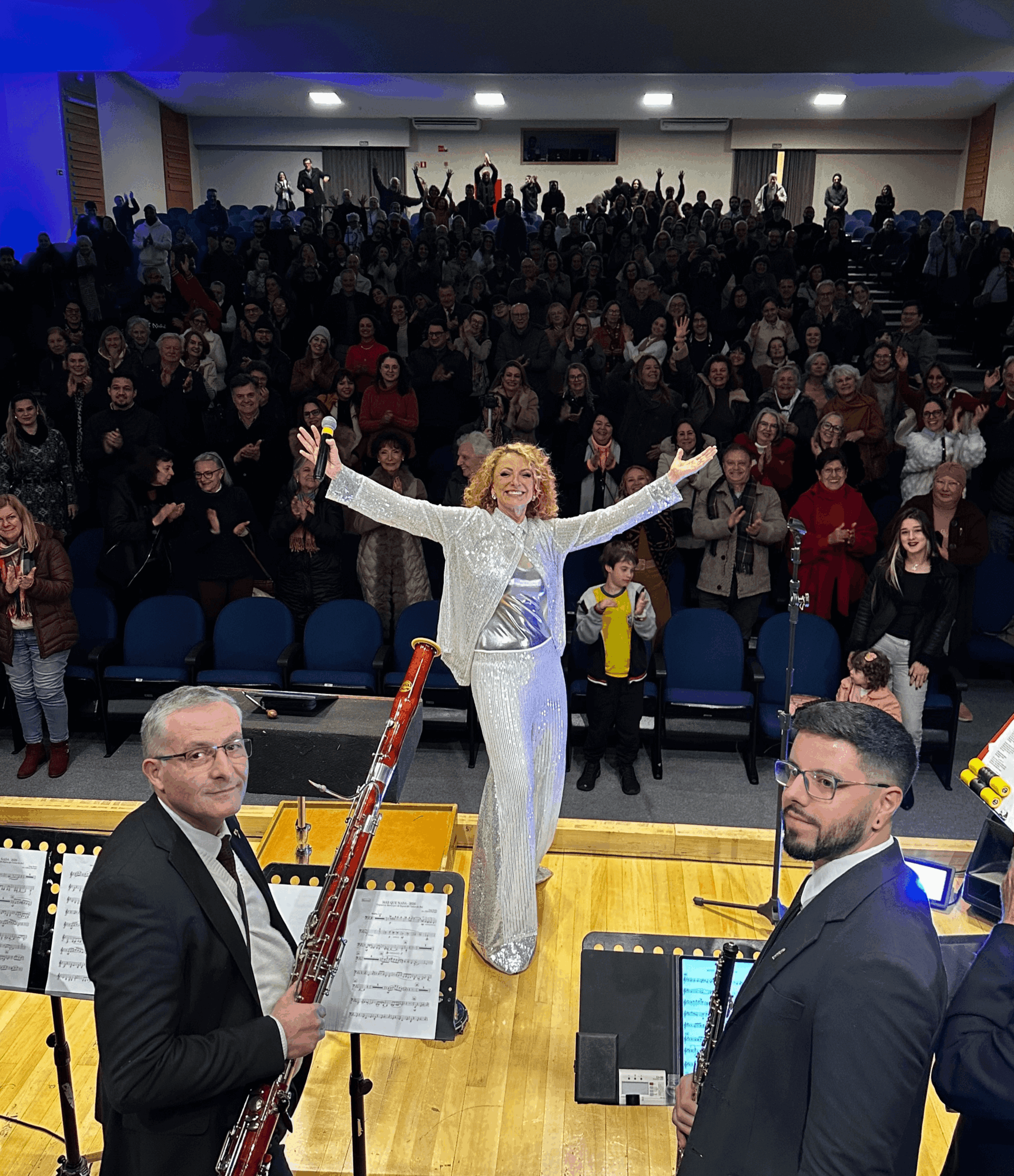 A woman in a silver outfit stands on stage with arms raised, facing an audience in a large hall, two men with musical instruments in foreground.