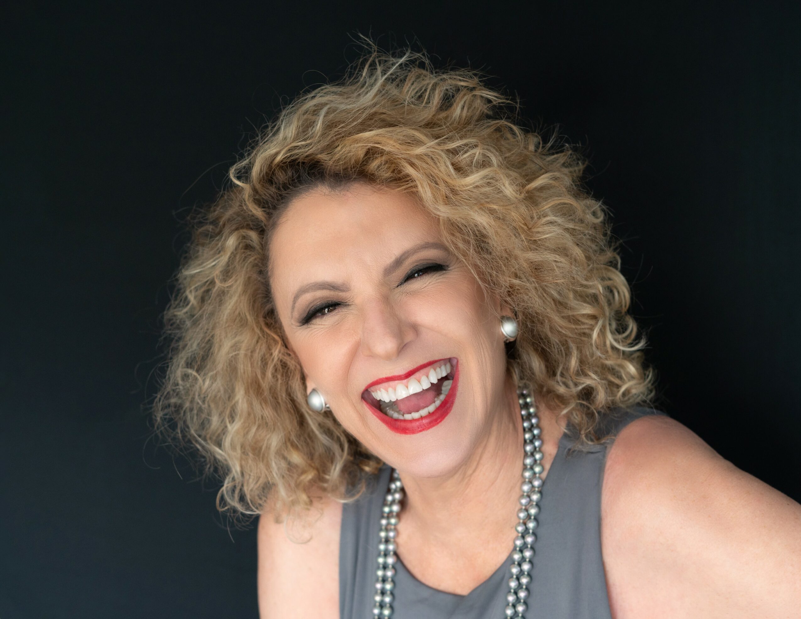 Woman with curly blonde hair smiling and winking, wearing a gray top and pearl necklace.
