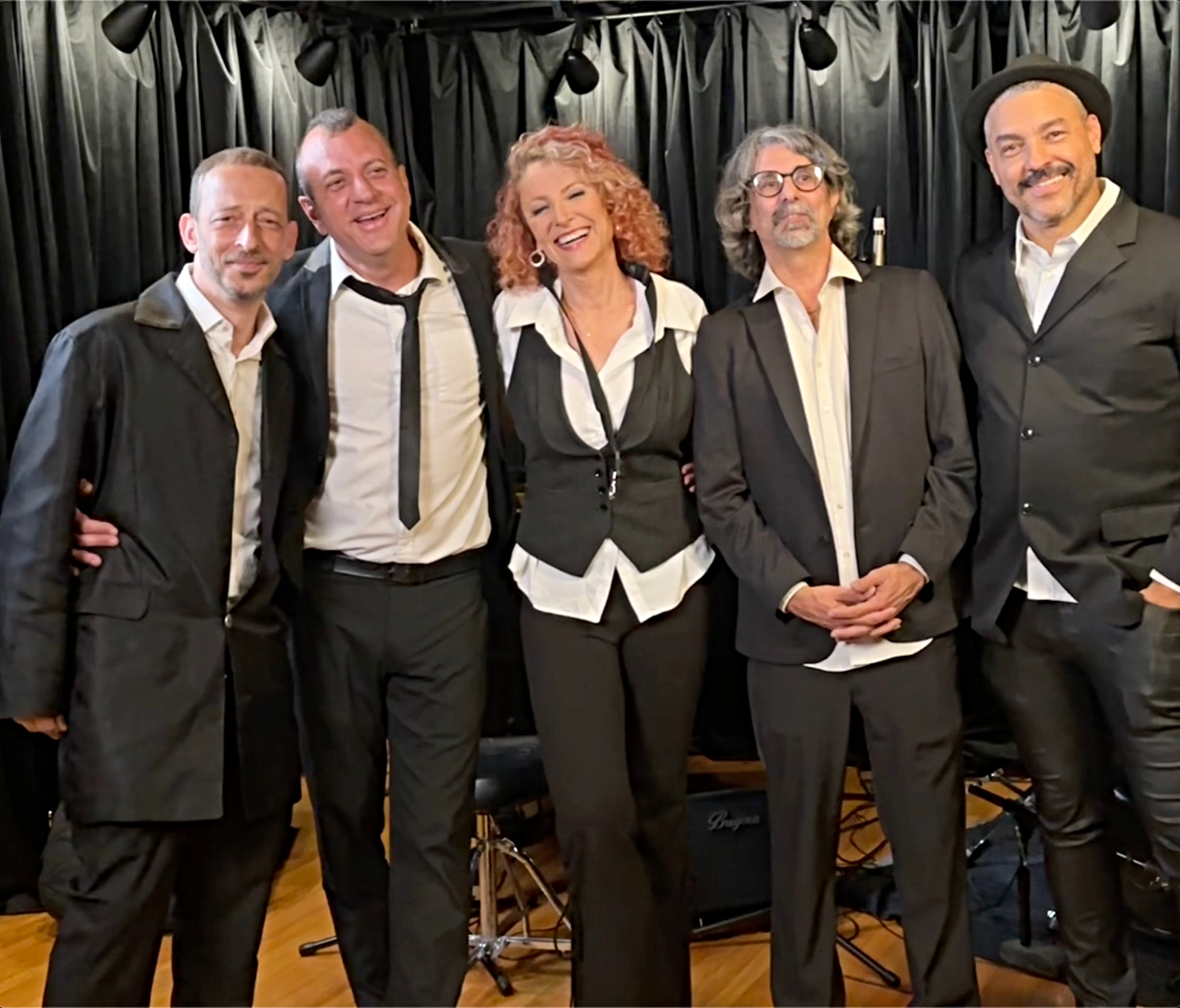 Five people standing together on stage, smiling, with a black curtain background, dressed in formal attire.