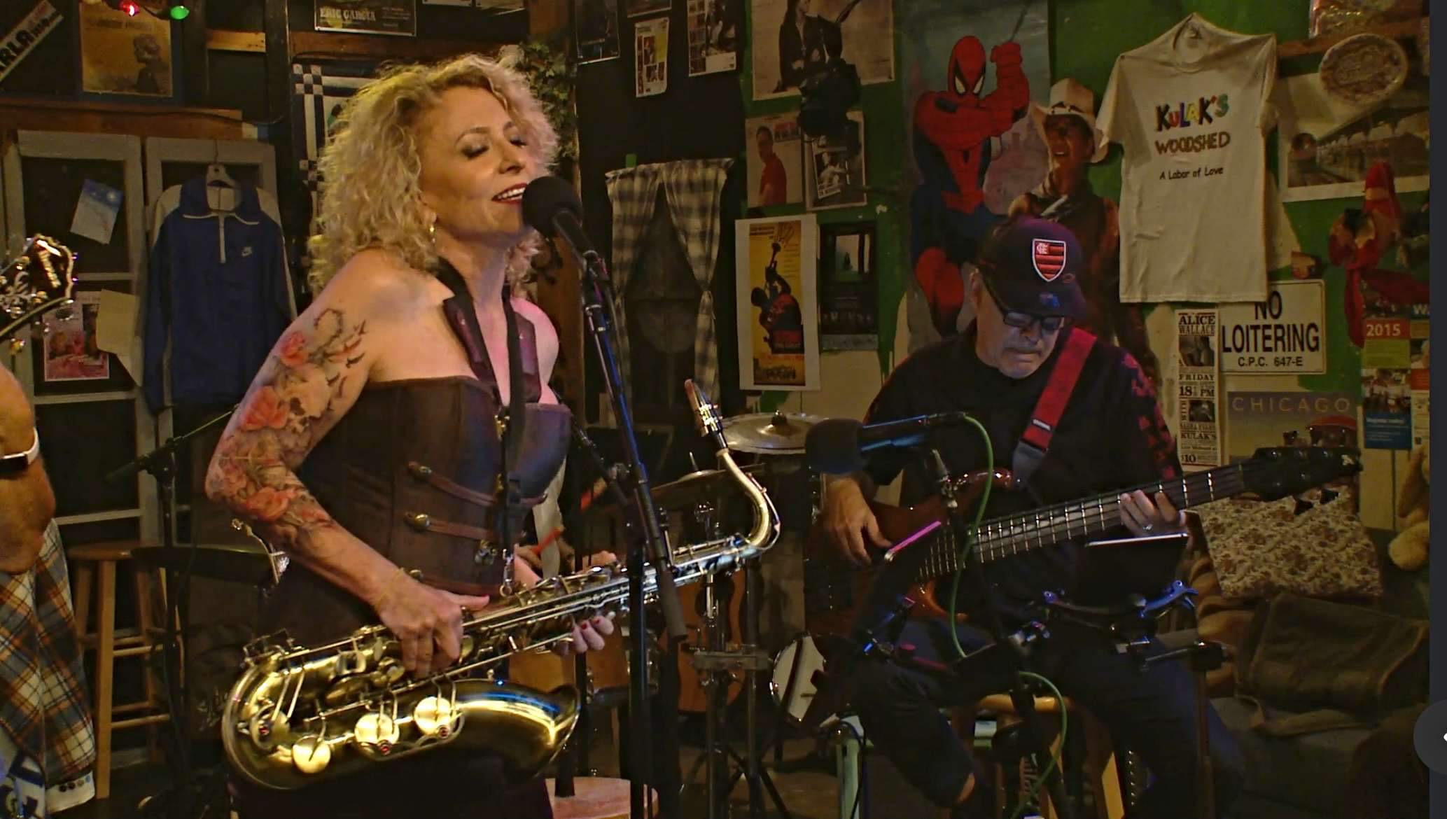 Two musicians perform on stage, one singing with a saxophone, the other playing guitar, in a colorful room with posters.