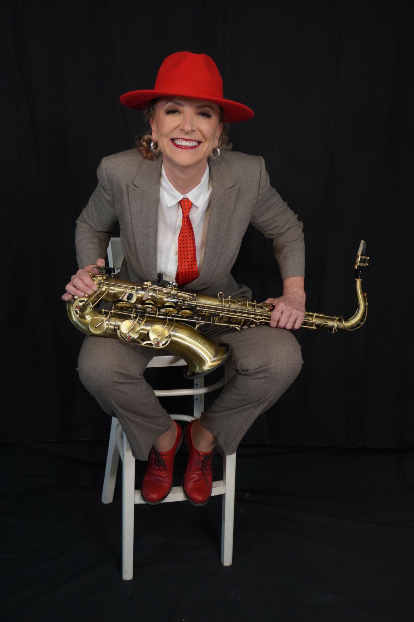 Person in red hat and gray suit holding a saxophone, sitting on a black background.
