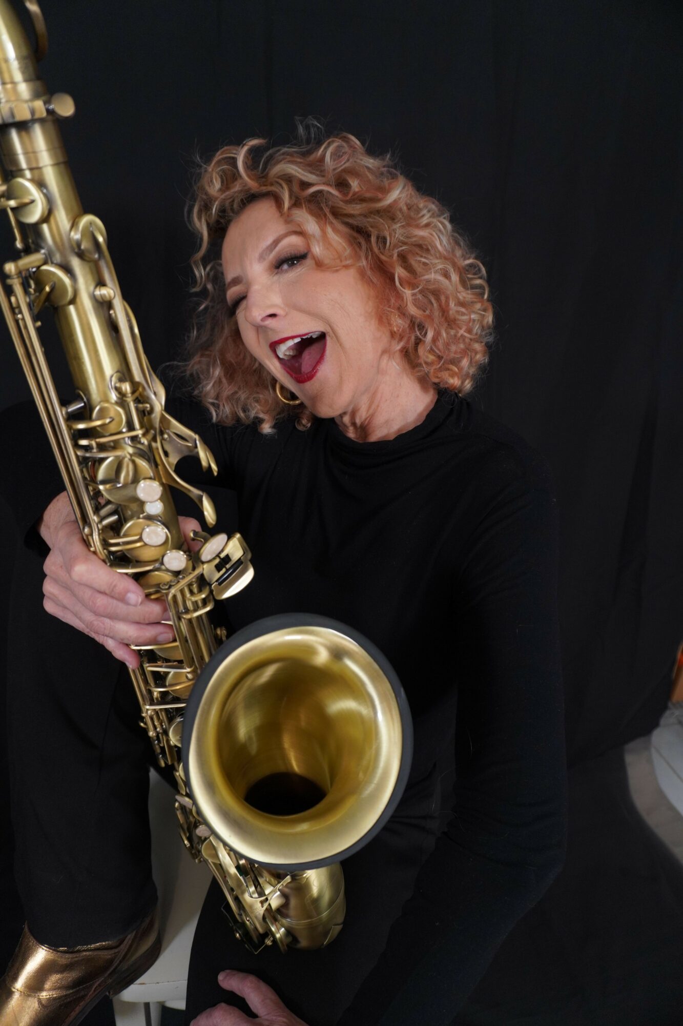 Woman with curly hair playing a saxophone, smiling with eyes closed, against a dark background.