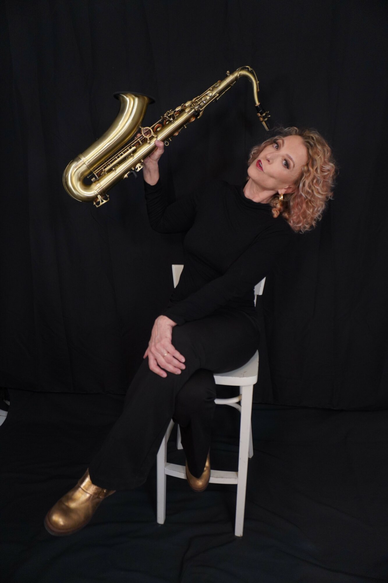 Person lying on a chair holding a saxophone, with a black background, wearing black clothing and gold shoes.