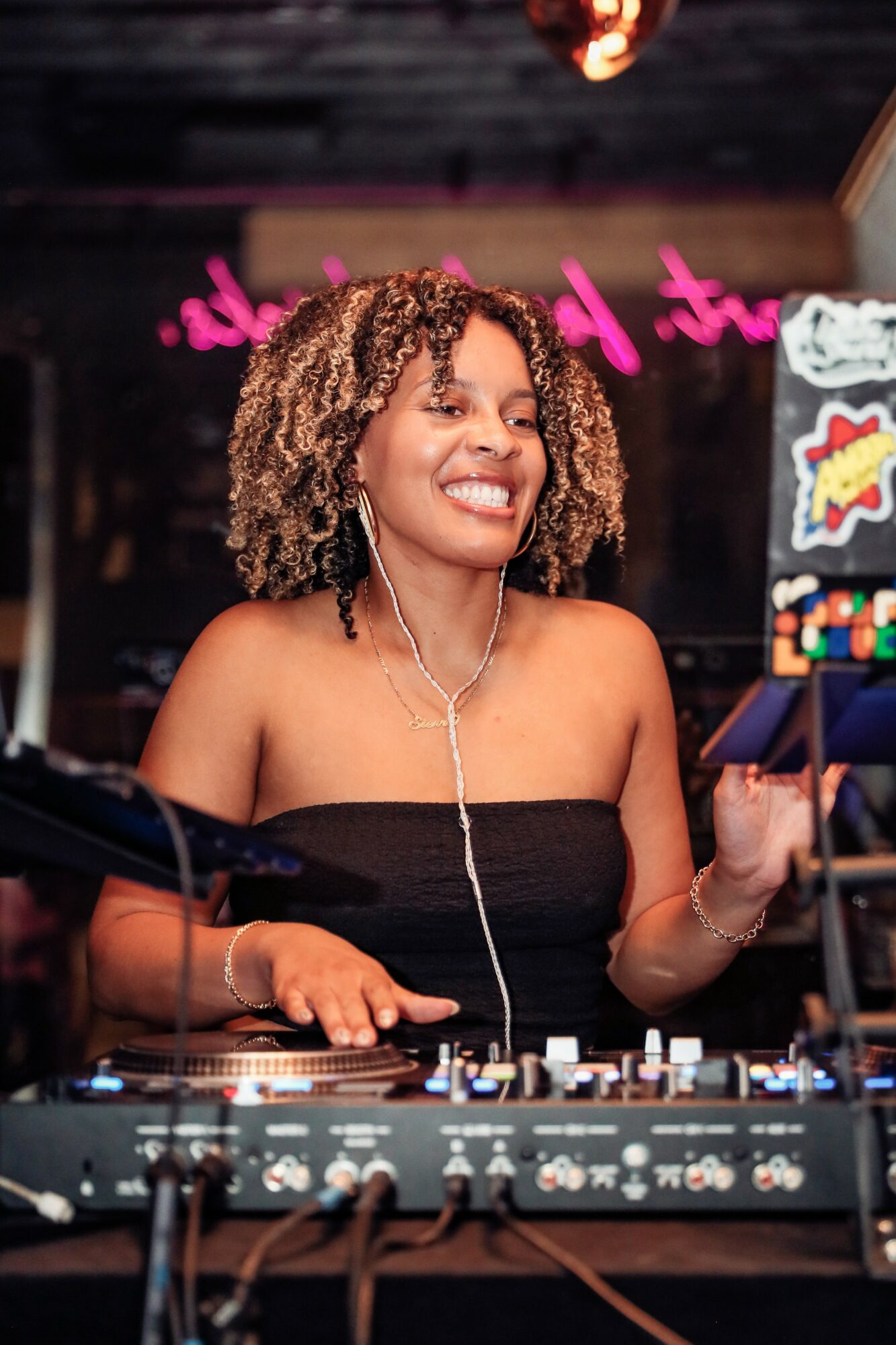Woman with curly hair smiling while DJing at a club or event, wearing a strapless top and headphones, with colorful signs in background.