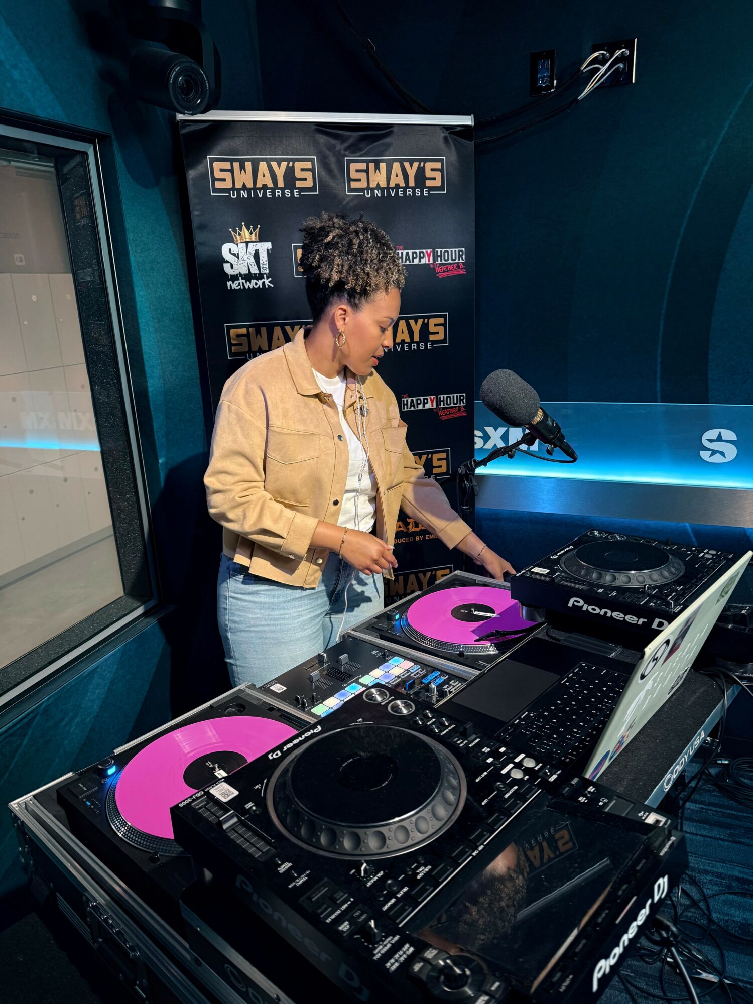 Woman DJ with curly hair wearing a beige jacket and white shirt, operating DJ equipment with pink turntables, in a studio.
