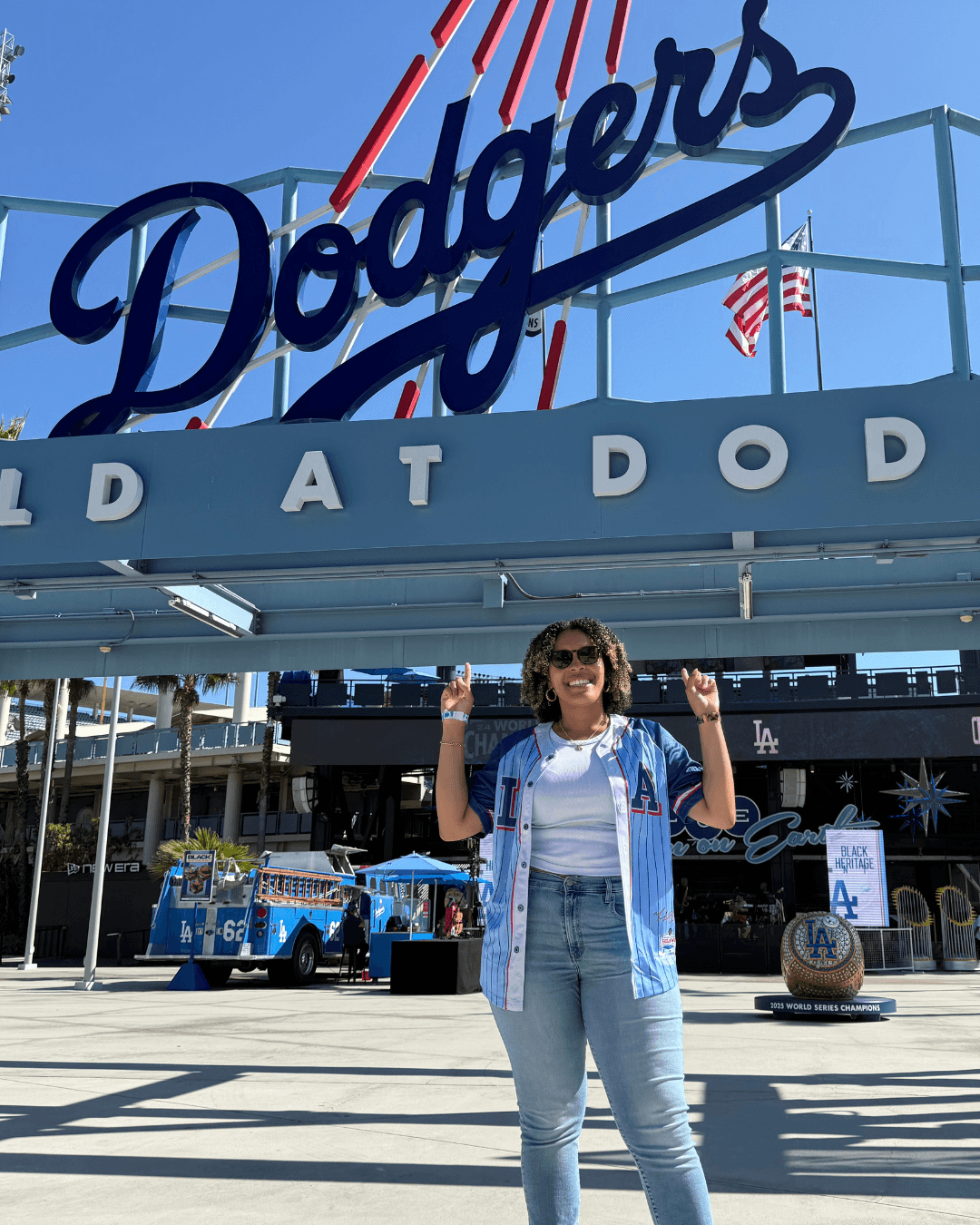 Woman standing in front of Dodger Stadium entrance, smiling with arms raised, blue sky above, baseball-themed decorations around.