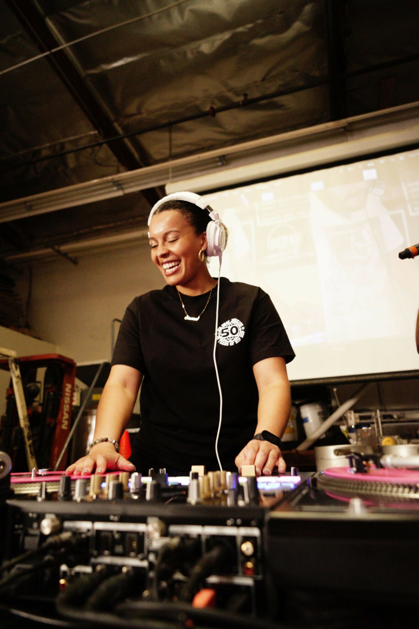 A woman DJ wearing headphones smiles while operating DJ equipment in a studio with a window behind her.