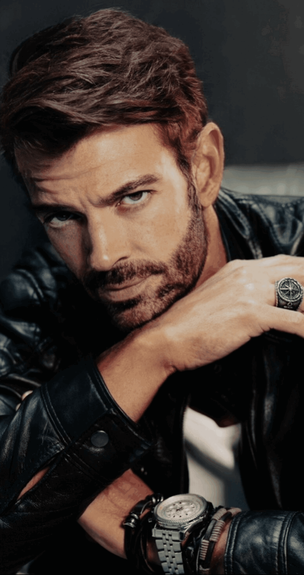 Man with brown hair and beard wearing a black leather jacket, resting his chin on his hand, looking at the camera.