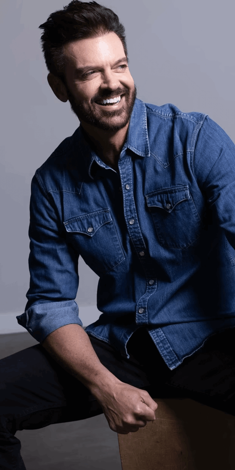 Man with dark hair and beard smiling, wearing a denim shirt, sitting against a plain background.