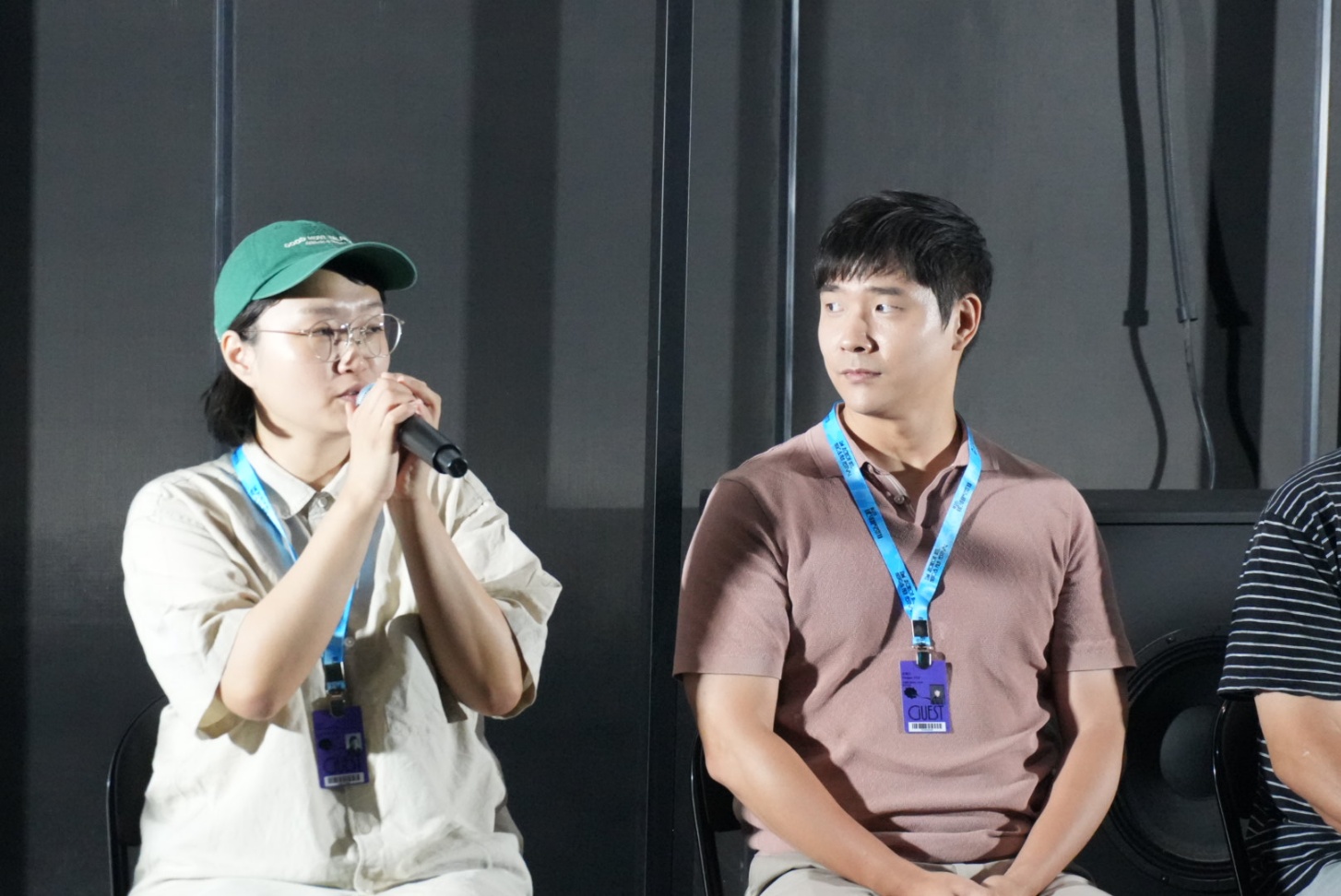 Two people standing indoors, one speaking into a microphone, the other listening, both wearing lanyards, dark background.