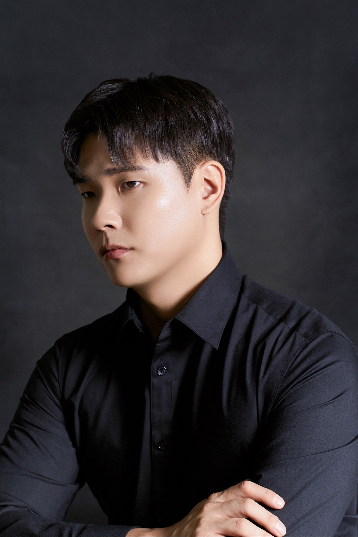 Young man with short dark hair wearing a black shirt, looking to the side against a dark background.