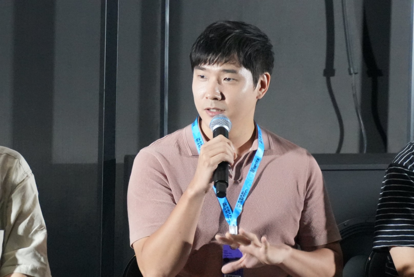 Young man speaking into a microphone, wearing a brown shirt and blue lanyard, seated indoors.