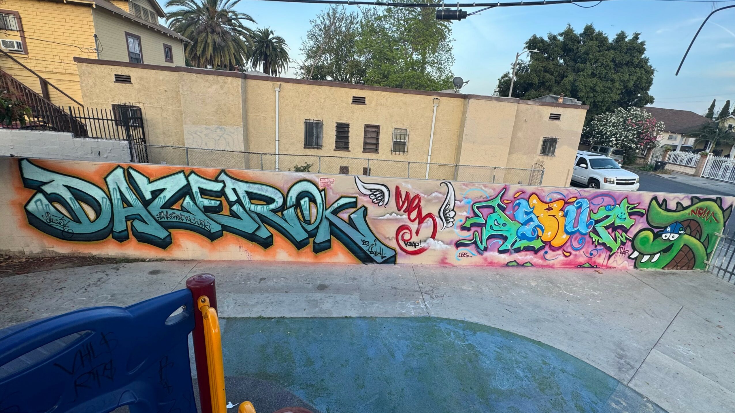 Graffiti art on a wall with colorful letters and designs, in an outdoor urban setting with trees and buildings in the background.