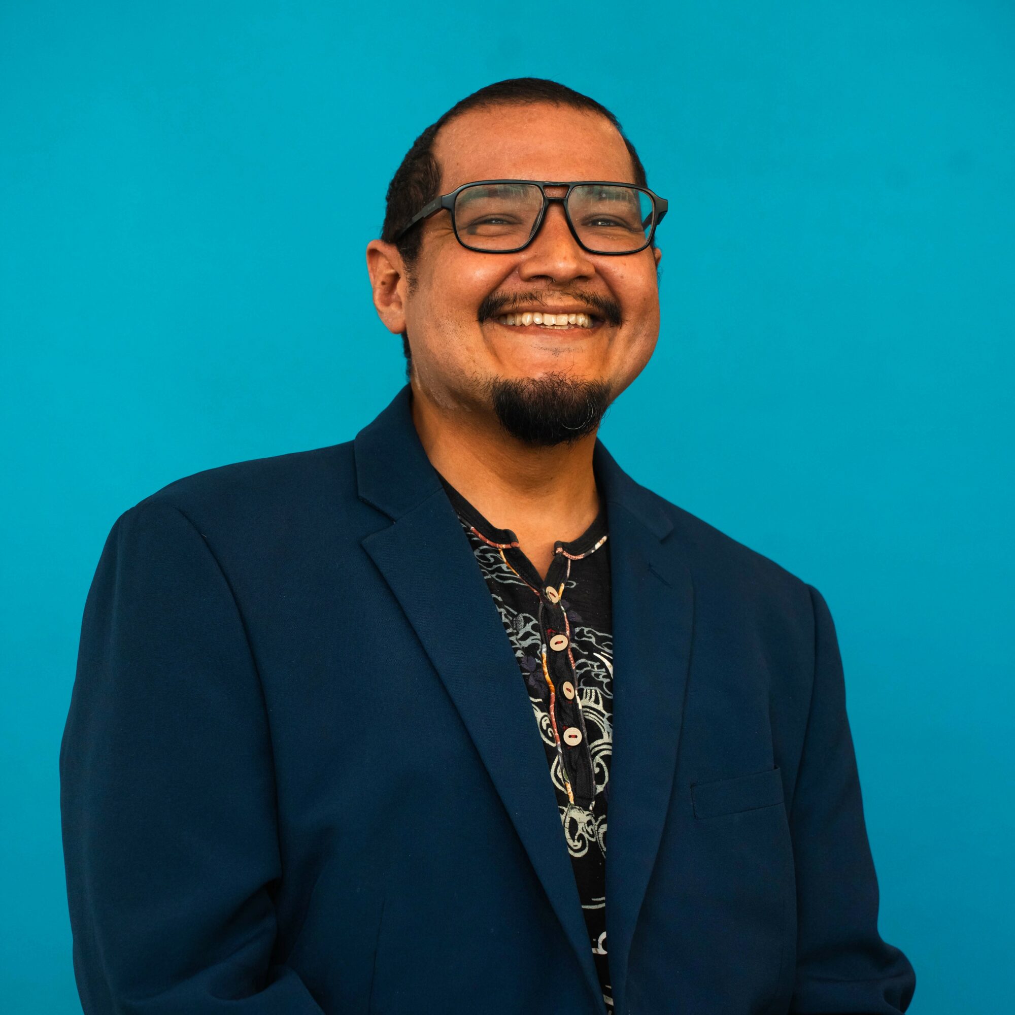 Smiling man with glasses and a beard wearing a dark blazer and patterned shirt against a blue background.
