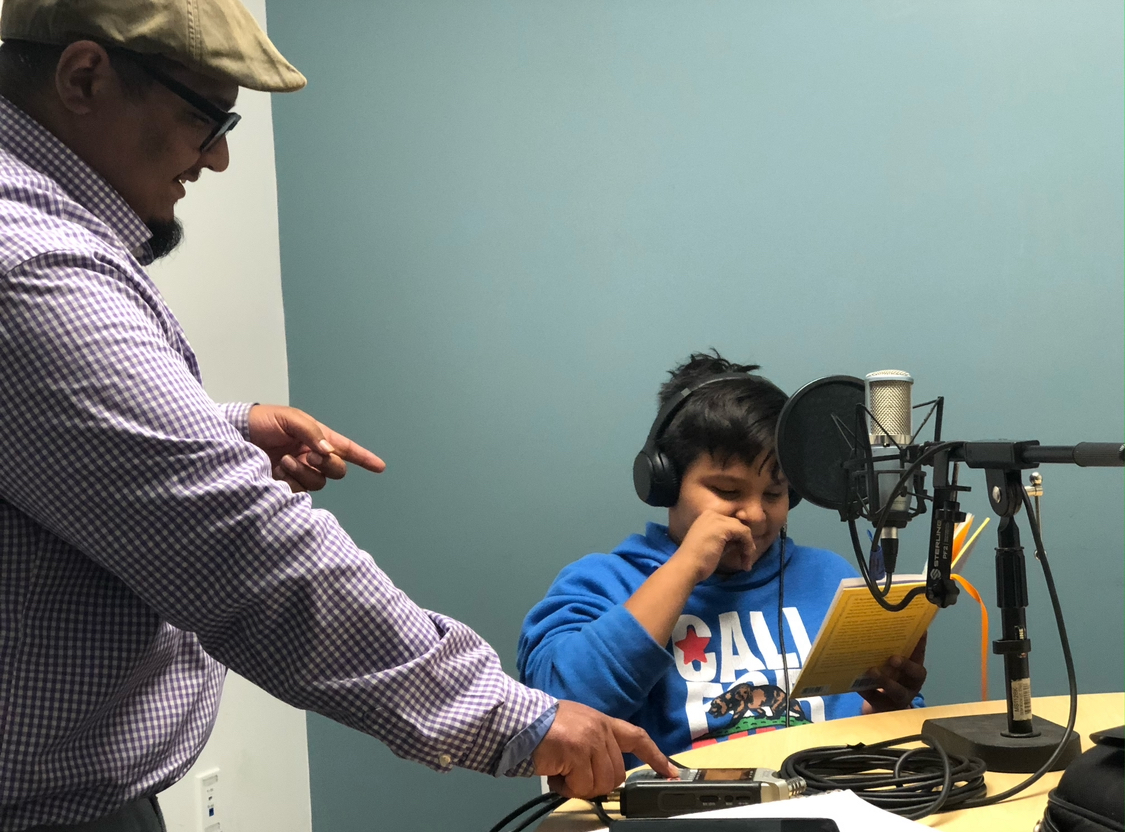 A person in a checkered shirt and cap points at a young boy in a blue hoodie recording in a studio.