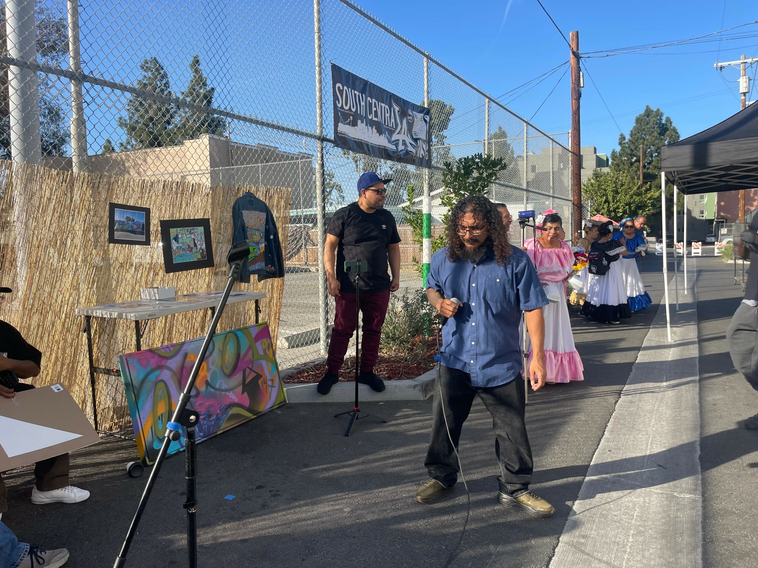 People walking along a sidewalk with outdoor art displays and a chain-link fence, sunny day, some children in colorful clothing.