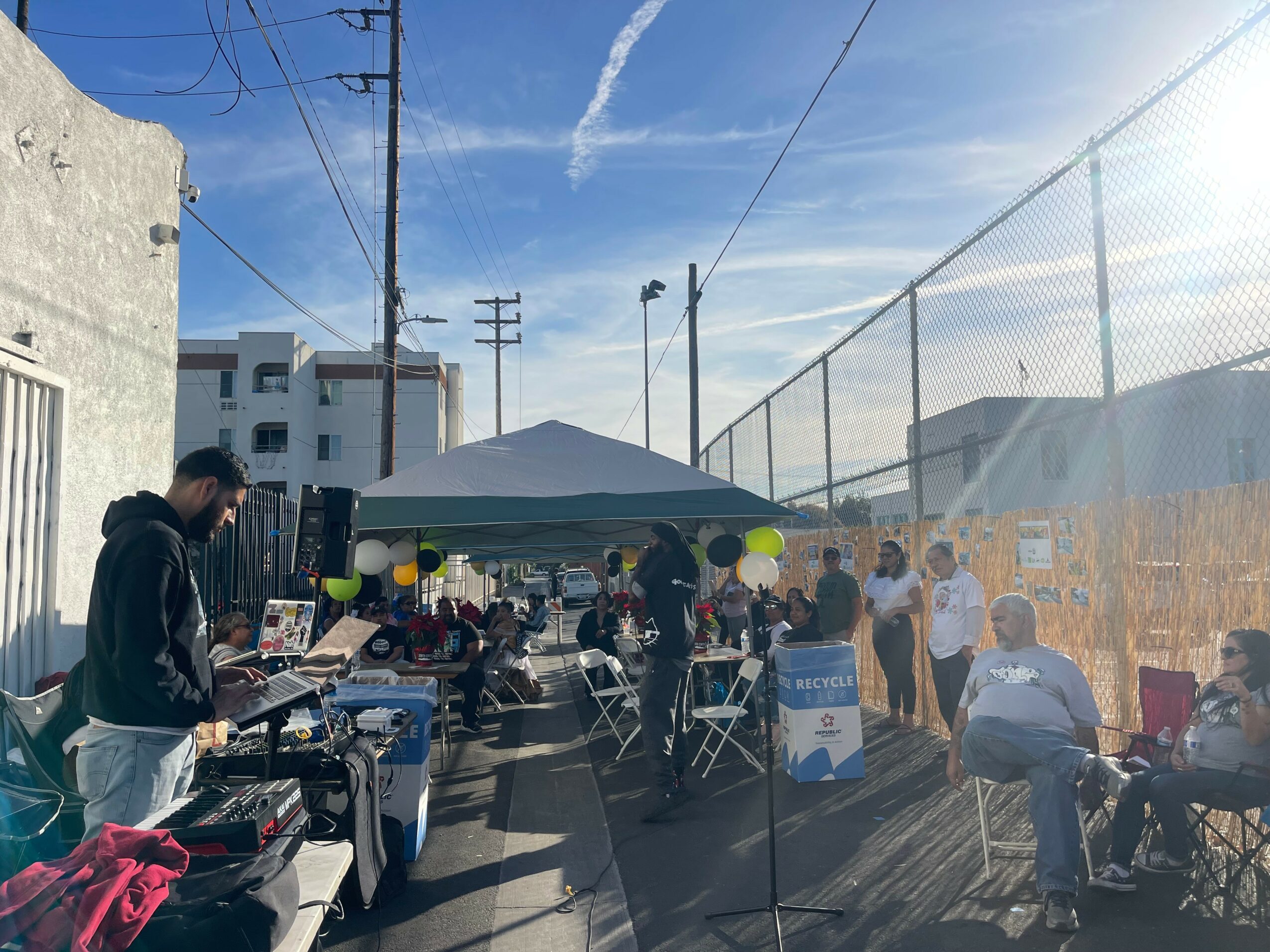 People gather under a tent and around tables outdoors on a sunny day, with a chain-link fence and buildings in background.