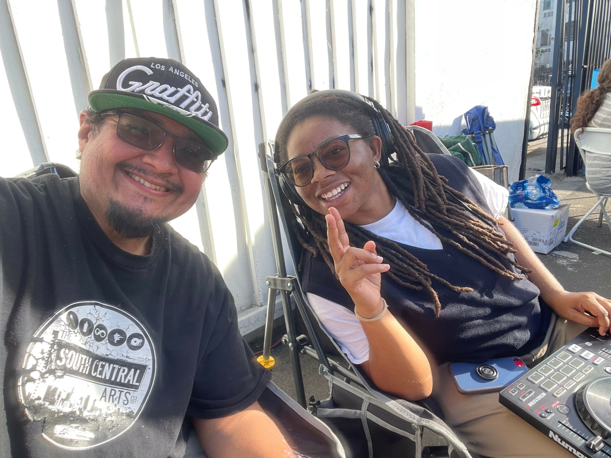 Two people smiling inside a vehicle, one wearing sunglasses and the other wearing a cap, with a bright background.