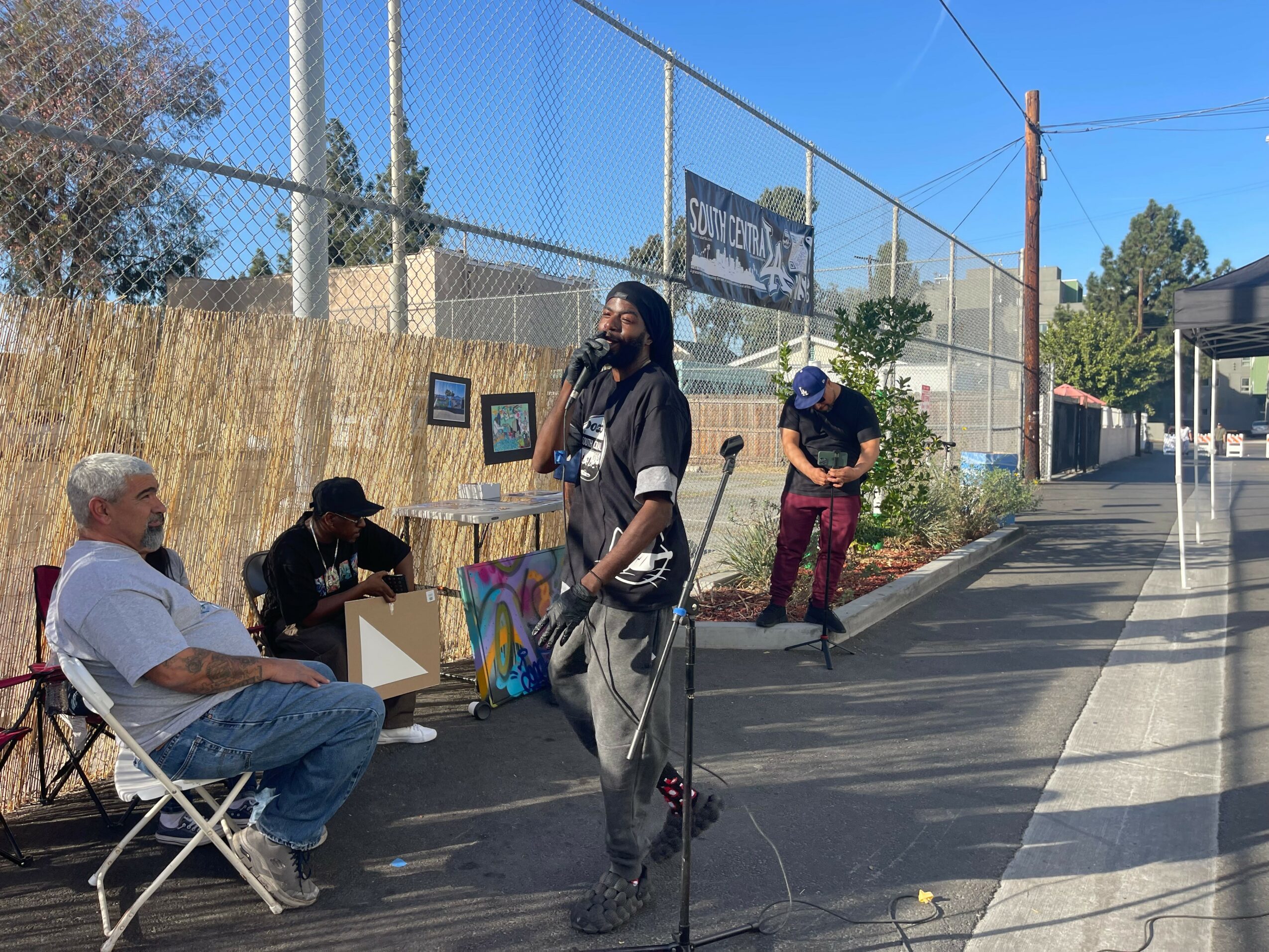 Four people are near a chain-link fence, with one person sitting and three standing, some with equipment, outdoors on a sunny day.