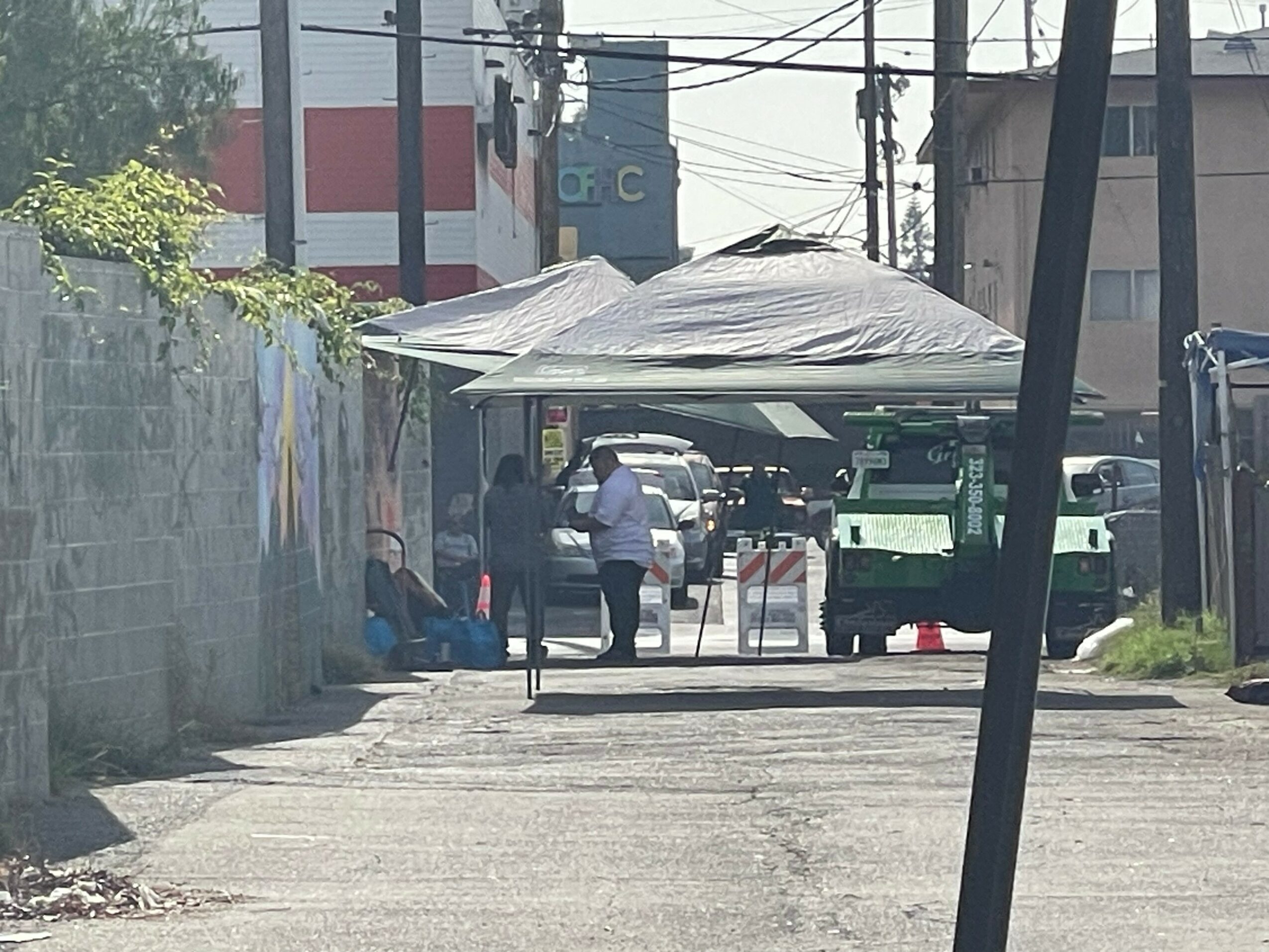 People standing under a canopy in an alleyway with vehicles and buildings in the background.