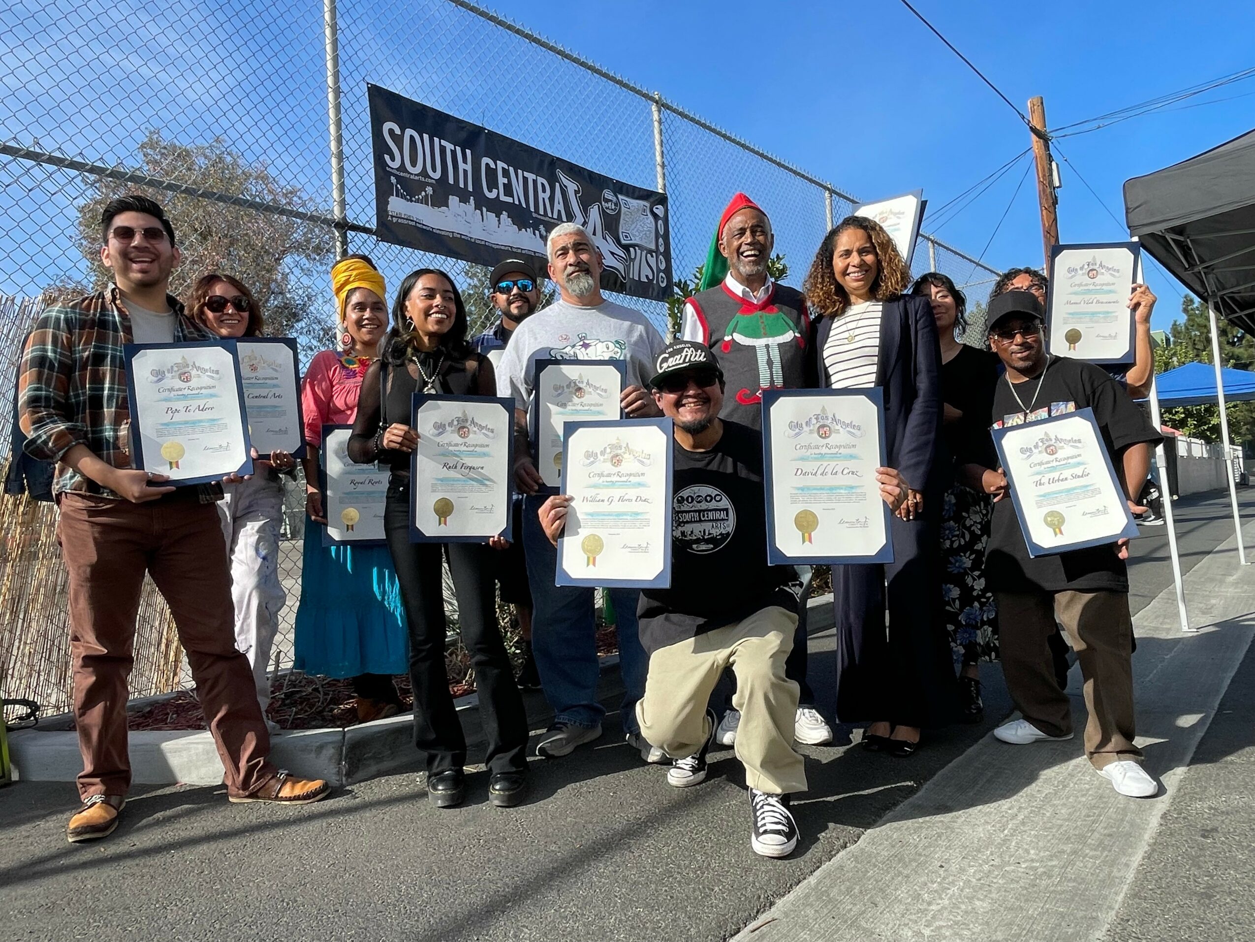 Group of ten people outdoors holding certificates, standing on a street with a chain-link fence and blue sky in background.