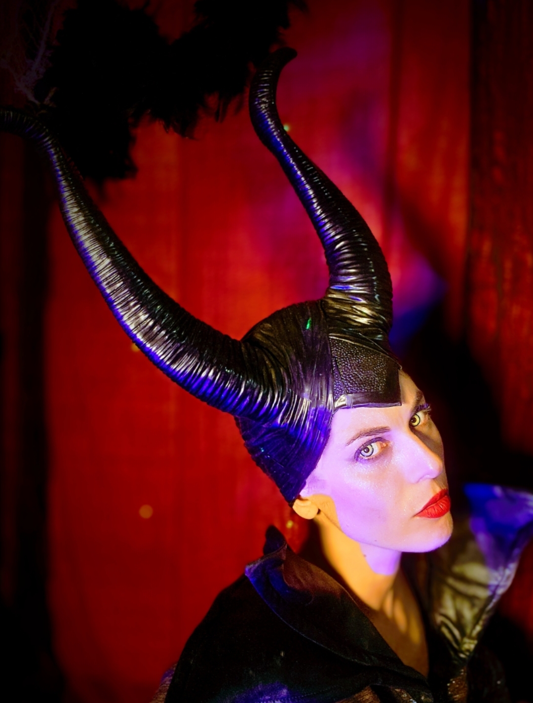 Person with large, curved, black horns and dramatic makeup, in a dark setting with red and purple lighting.
