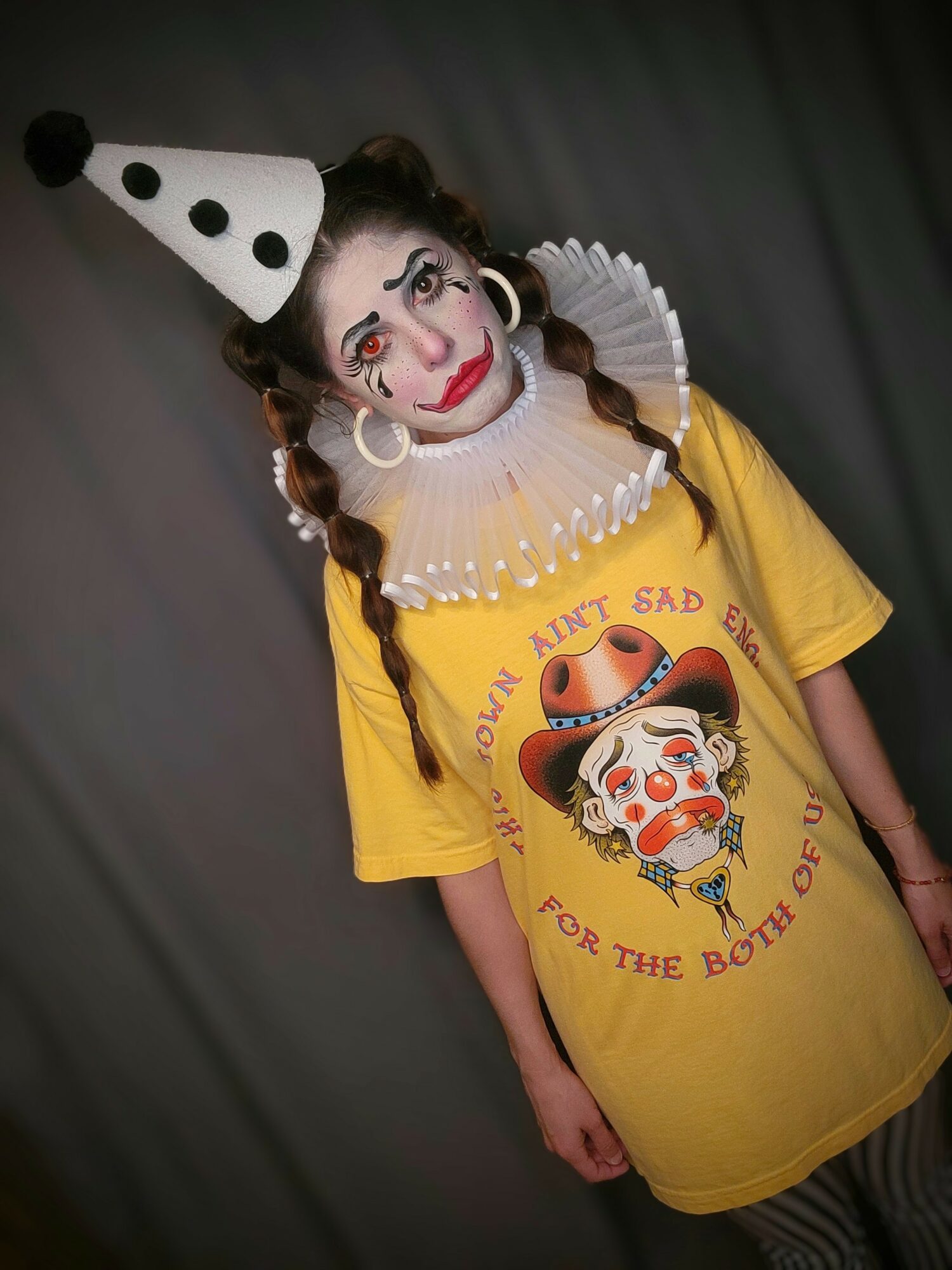 Child dressed as a clown with face paint, wearing a yellow T-shirt with a clown graphic, and a polka dot hat, standing against a dark background.