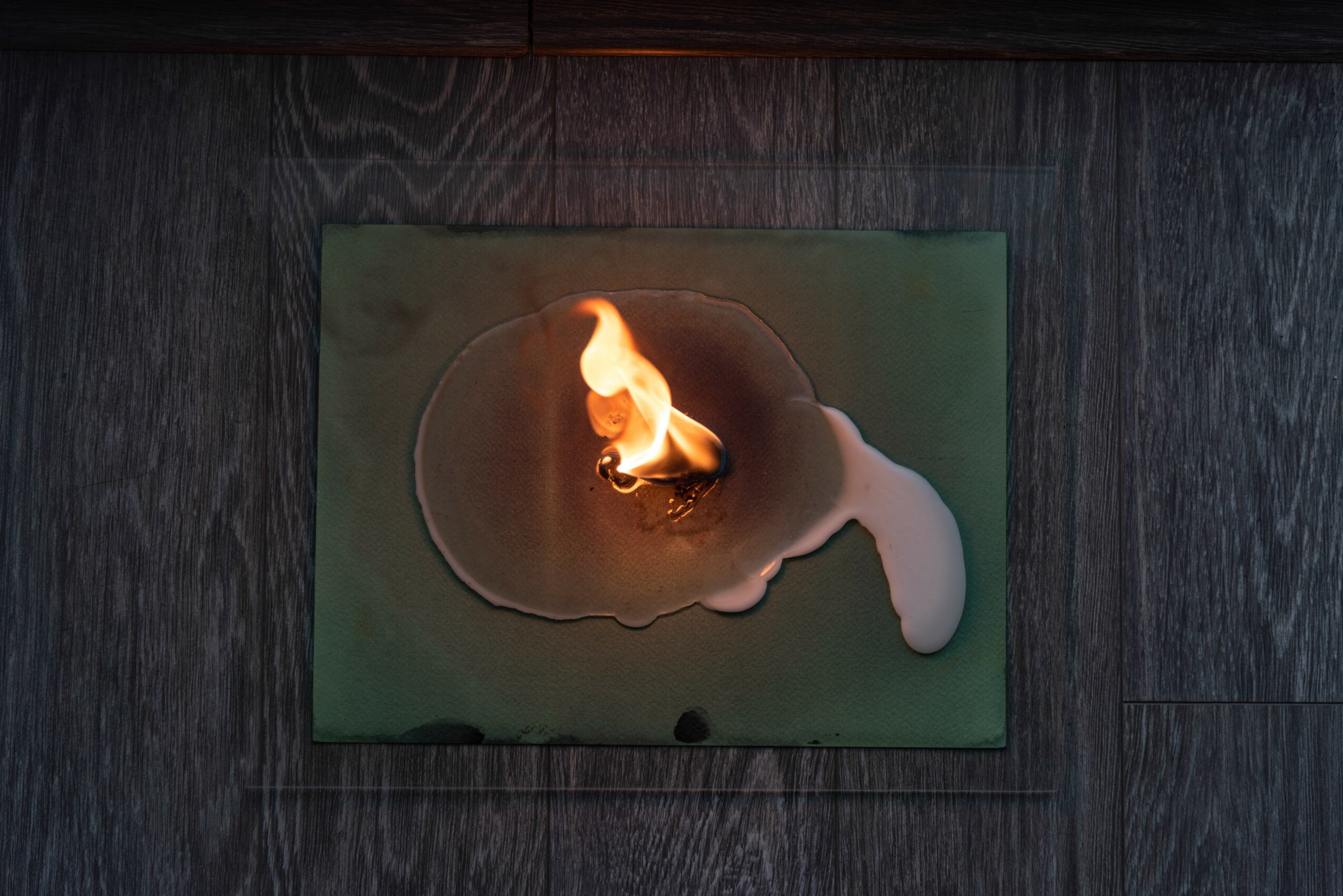 A candle flame burns on a green surface with wax melting and pooling around it.
