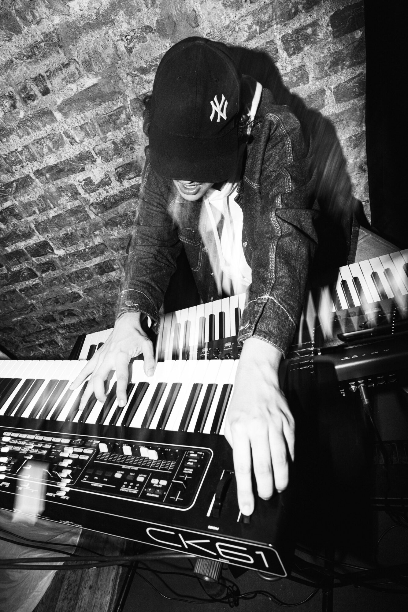 Person playing keyboard, wearing a cap, with hands on keys, against a brick wall background.