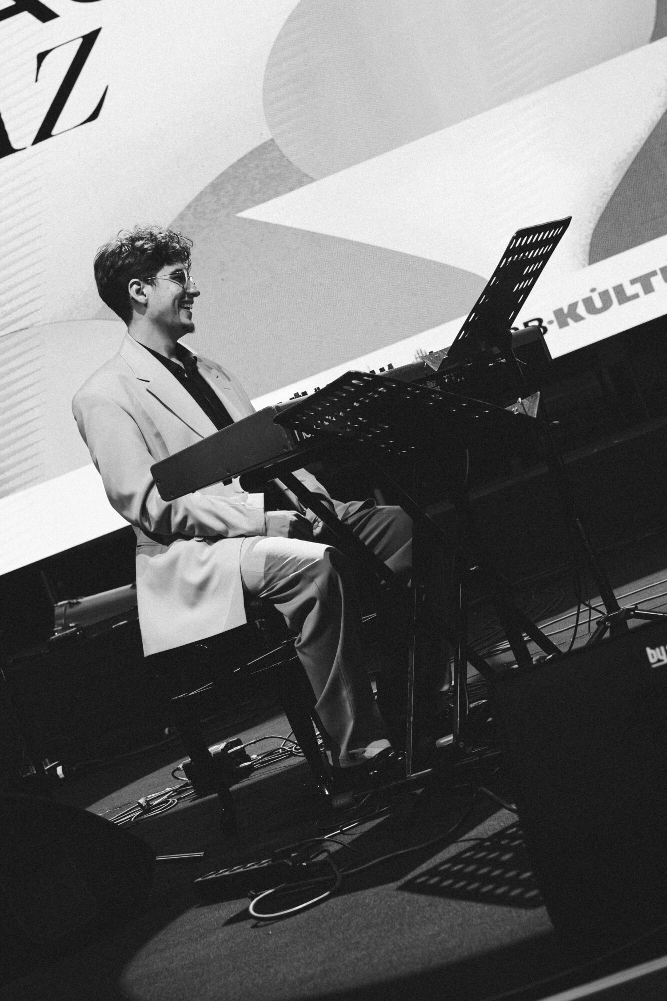 Person playing keyboard on stage with a large screen in the background, in black and white.
