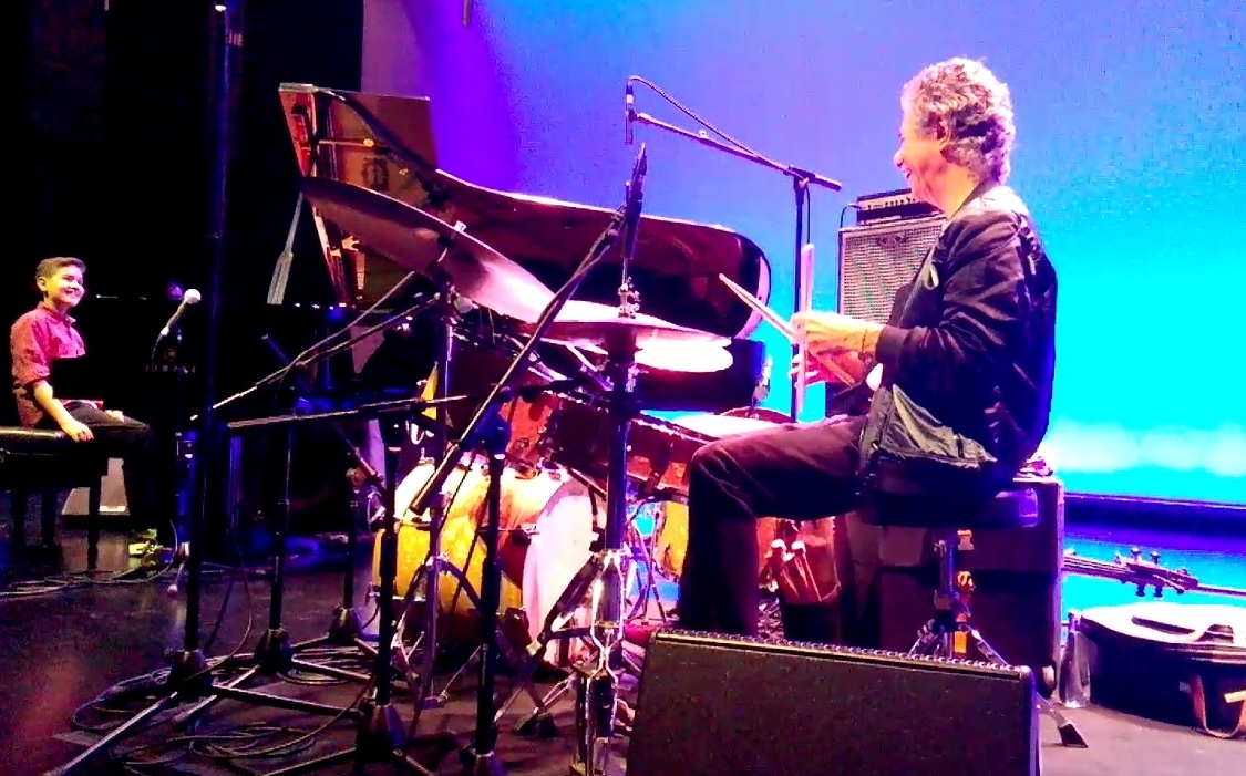 Three musicians perform on stage with a grand piano, drums, and a keyboard, illuminated by colorful stage lighting.