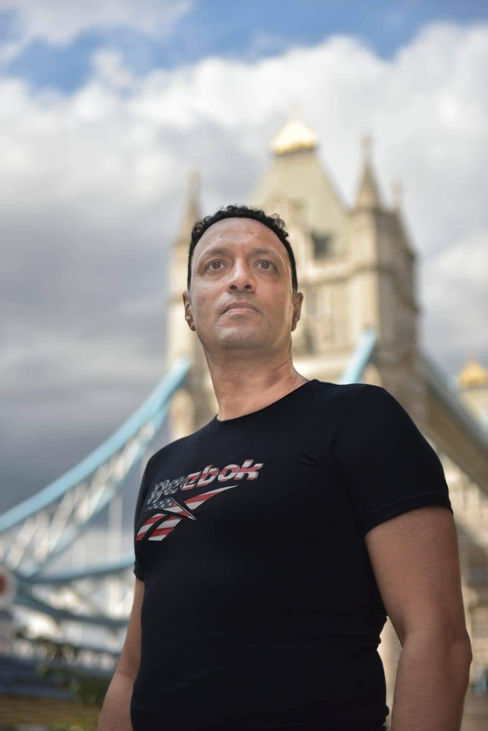 Man standing outdoors with a bridge and castle in the background, looking upward, cloudy sky above.