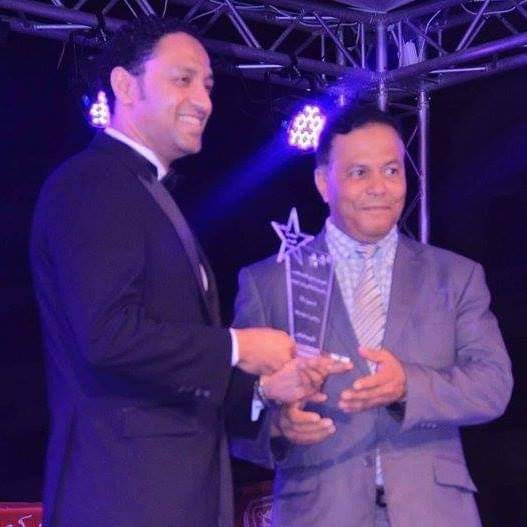 Two men in suits exchanging an award on stage with purple lighting and metal framework in background.