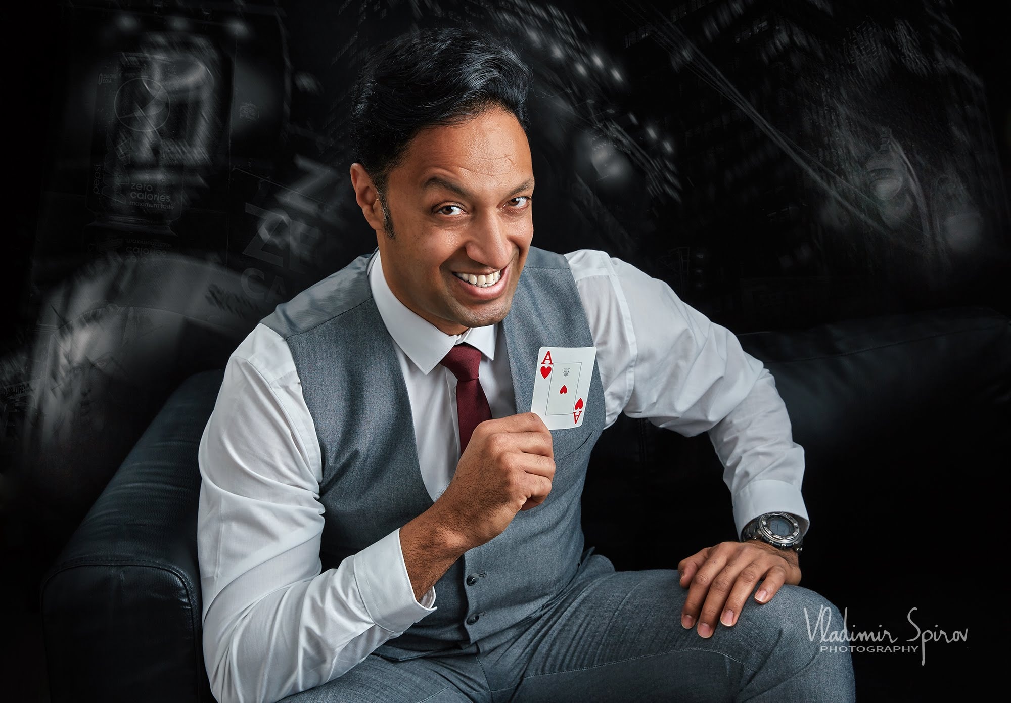 Man in suit holding playing card, smiling, sitting in dark room with abstract background.
