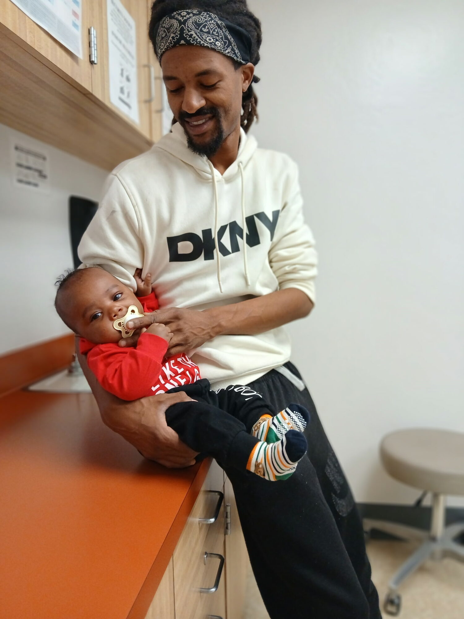 Adult holding a baby in a medical setting, both smiling, baby with a pacifier, adult wearing a DKNY hoodie.